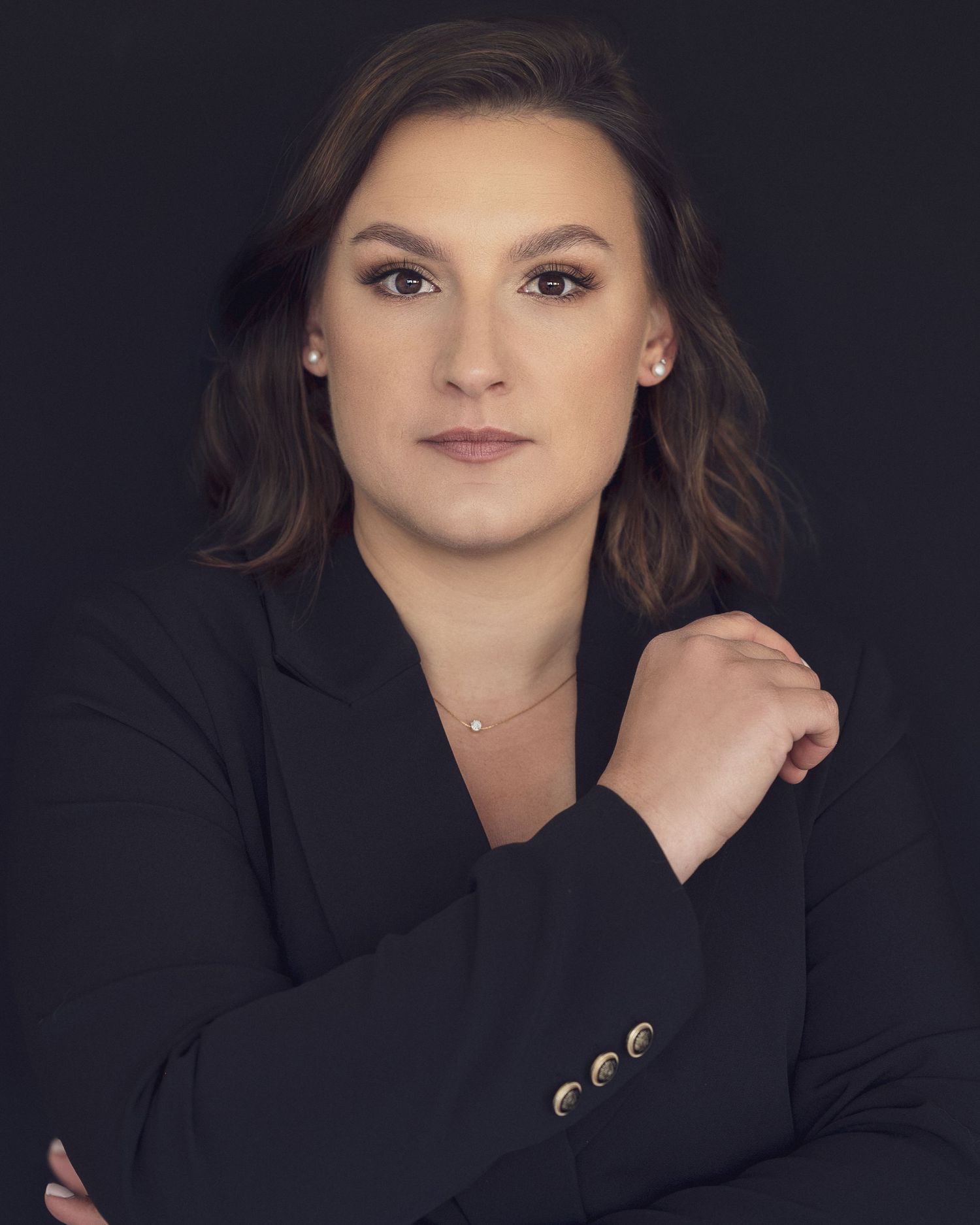 Professional woman in black blazer with arms crossed, posing confidently against dark background.