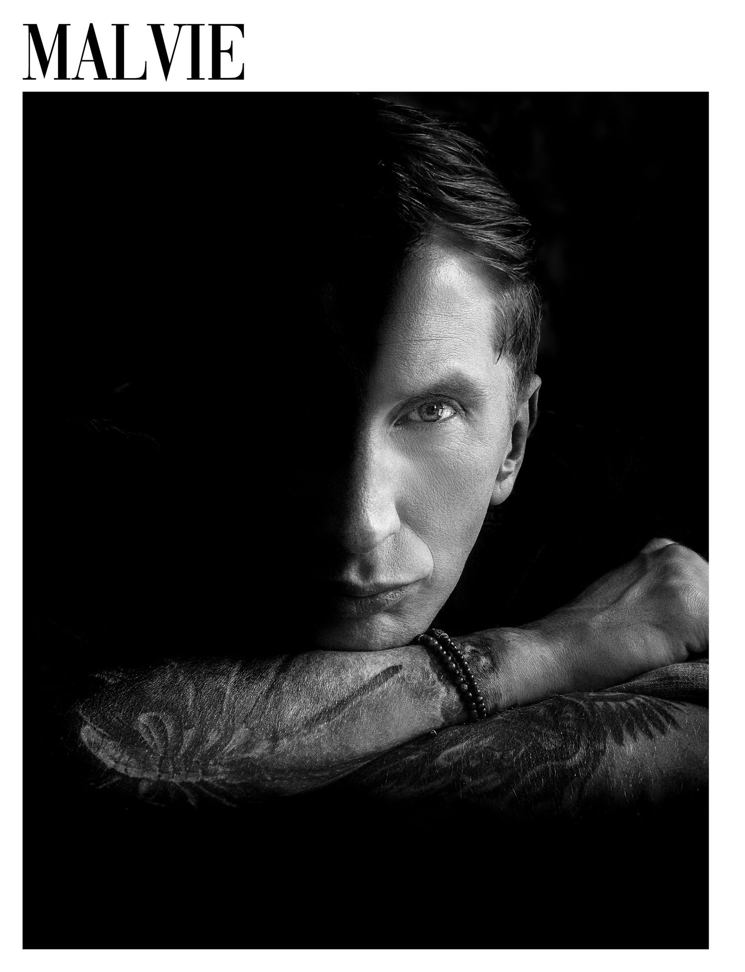 Black and white Malvie magazine portrait of a tattooed man resting his chin on crossed arms in dramatic lighting.