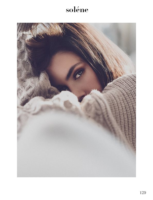 Close-up of a young woman with brown hair wrapped in cozy knit sweaters, gazing softly at the camera.