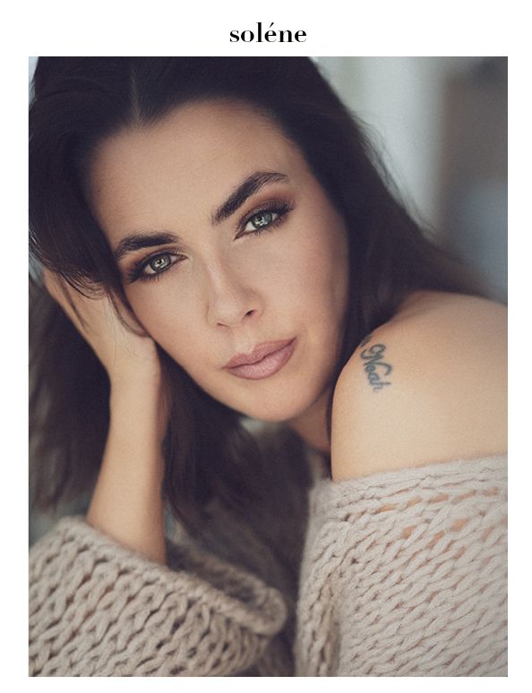 Close-up portrait of a brunette woman with smoky eye makeup wearing an off-shoulder knit sweater with a small tattoo.