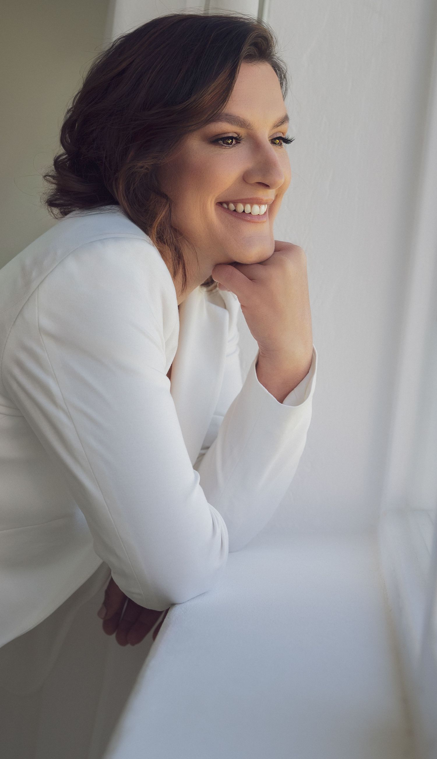 Smiling woman in white blazer leaning on windowsill with hand under chin, bright natural light portrait.