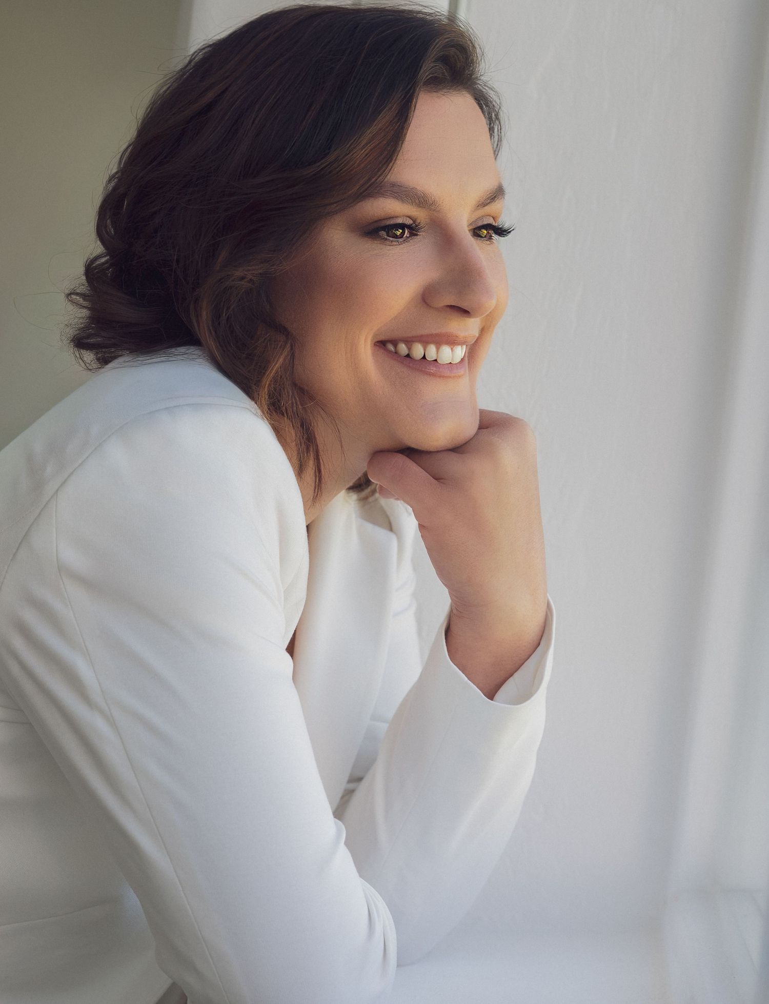 A smiling woman in a white blazer poses with hands clasped near her chin in a bright, elegant portrait photo.