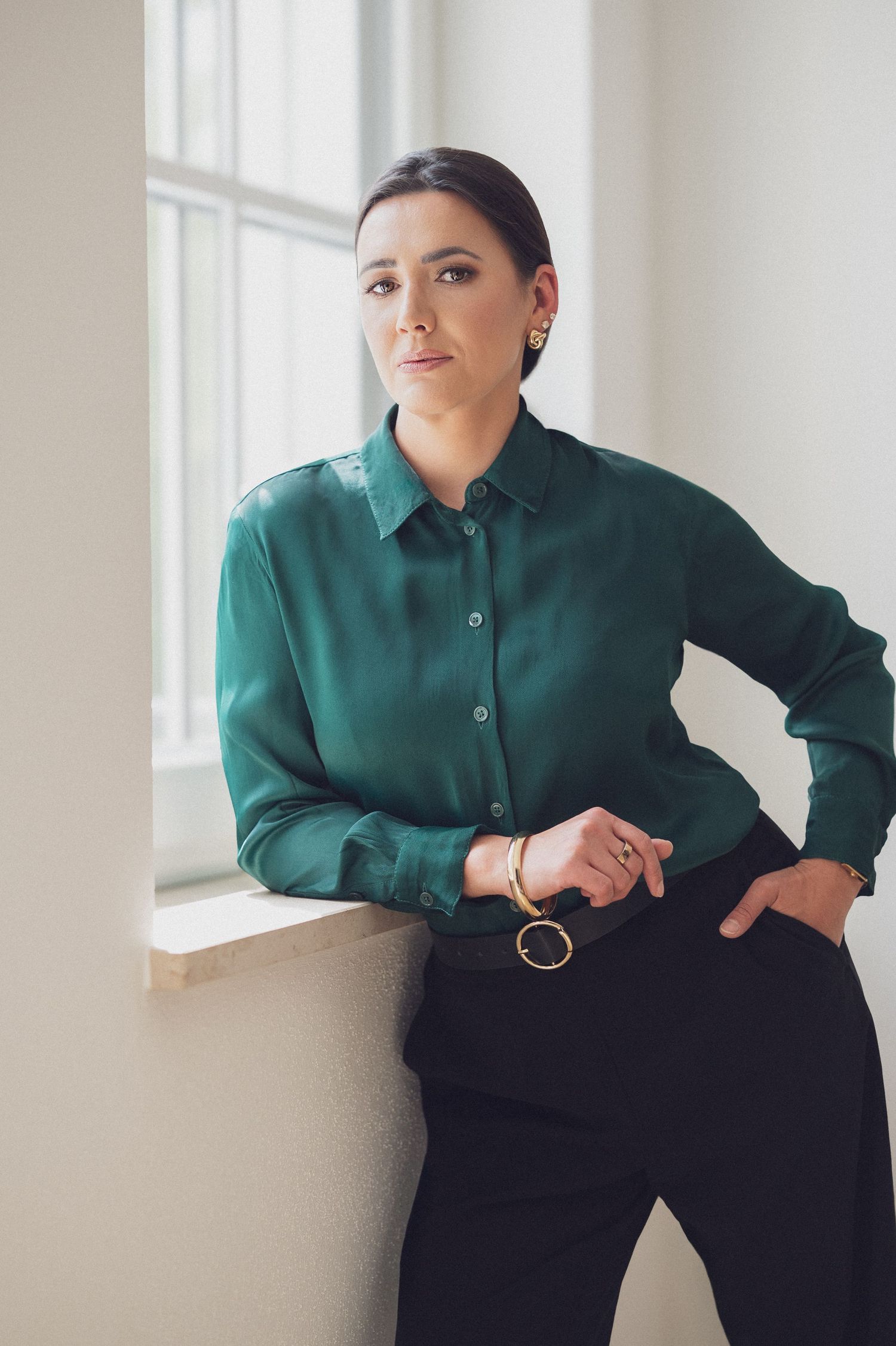 Woman in green silk blouse and black pants leaning against a window ledge, wearing gold jewelry.