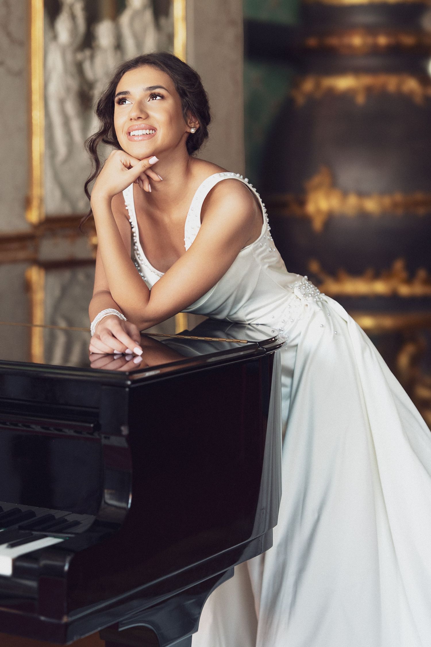 A smiling bride in a white gown leans on a grand piano, gazing upward in an elegant ornate room.