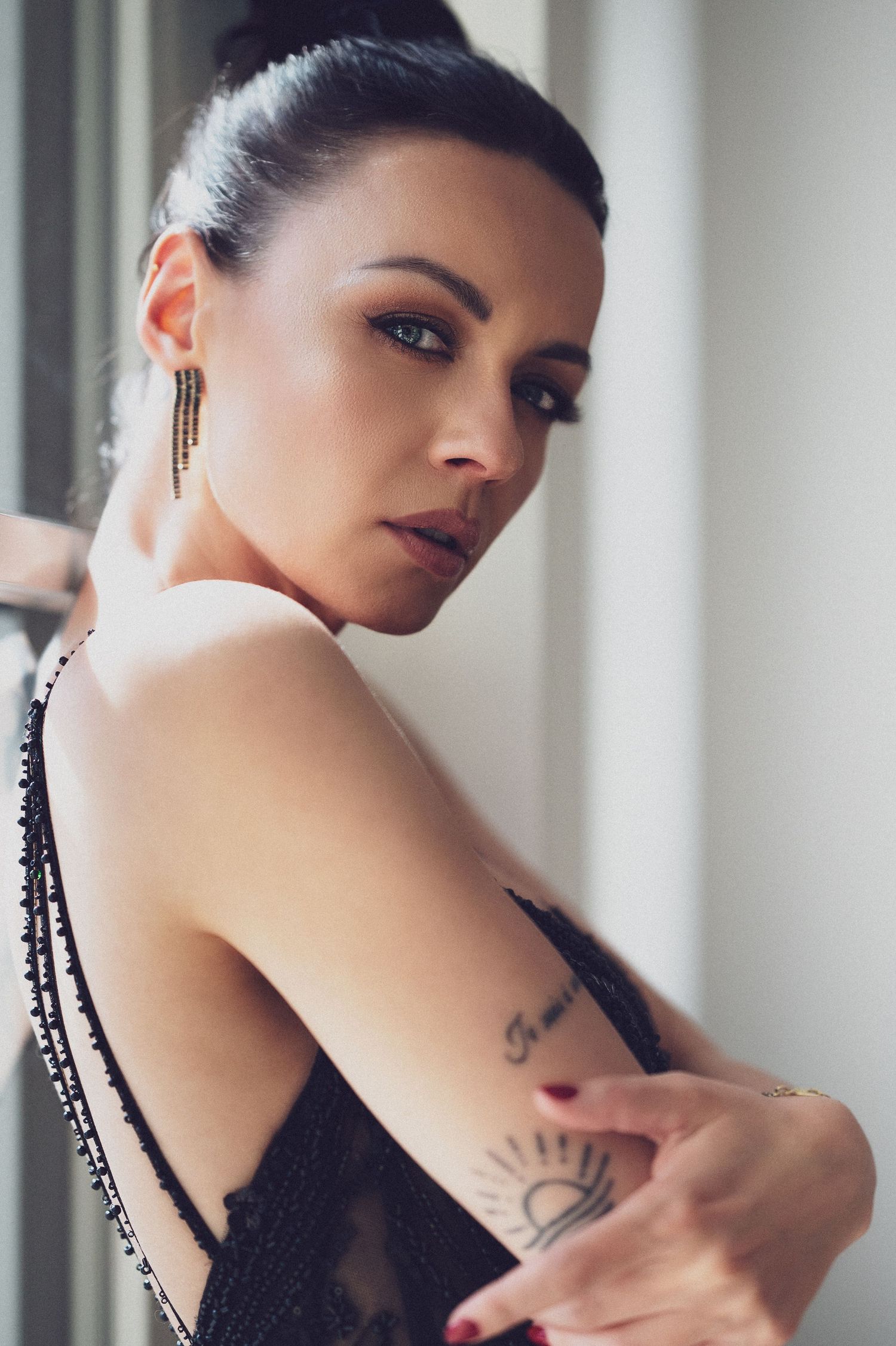 Woman with dark hair, gold earrings, and arm tattoo poses in black lace top near window.