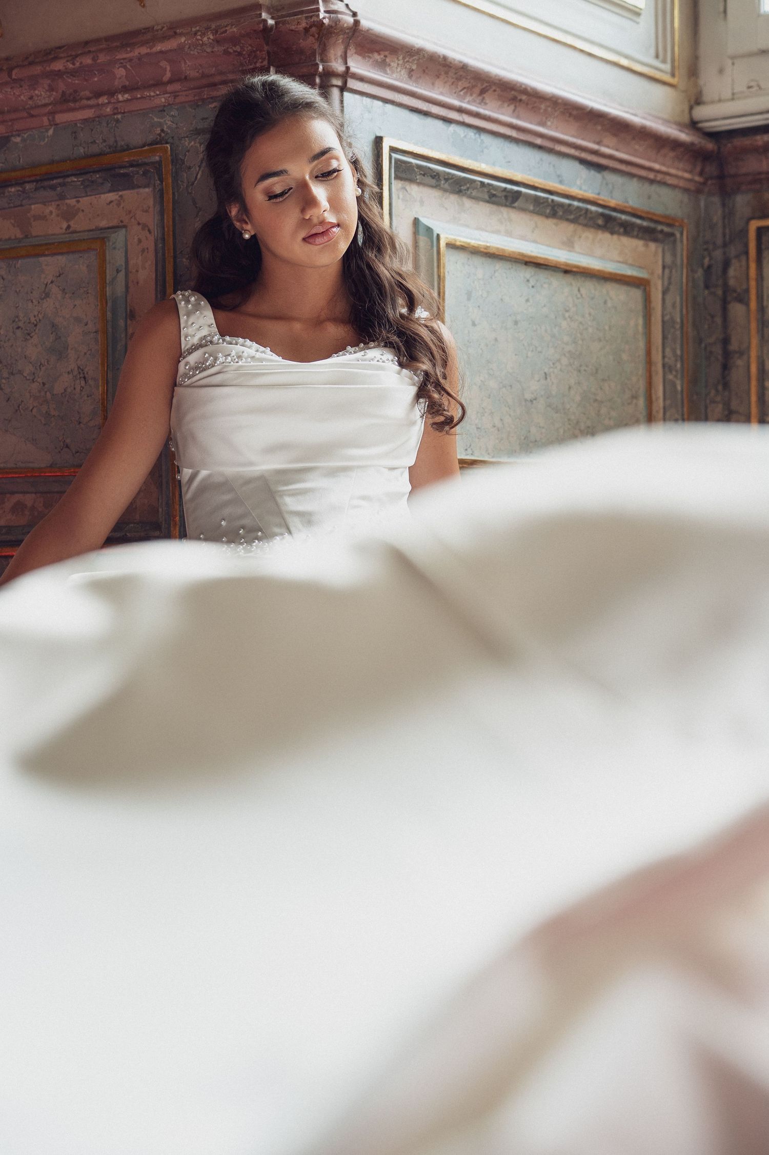Elegant bride in white wedding gown sitting gracefully against ornate vintage wall with flowing dress in foreground.
