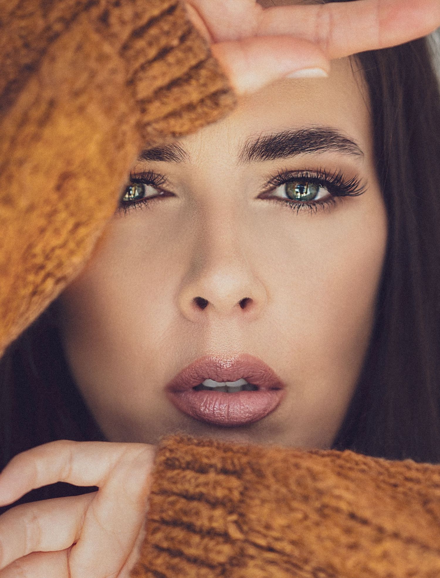 Close-up beauty portrait with dramatic lighting showing expressive eyes and makeup framed by brown textured fabric.
