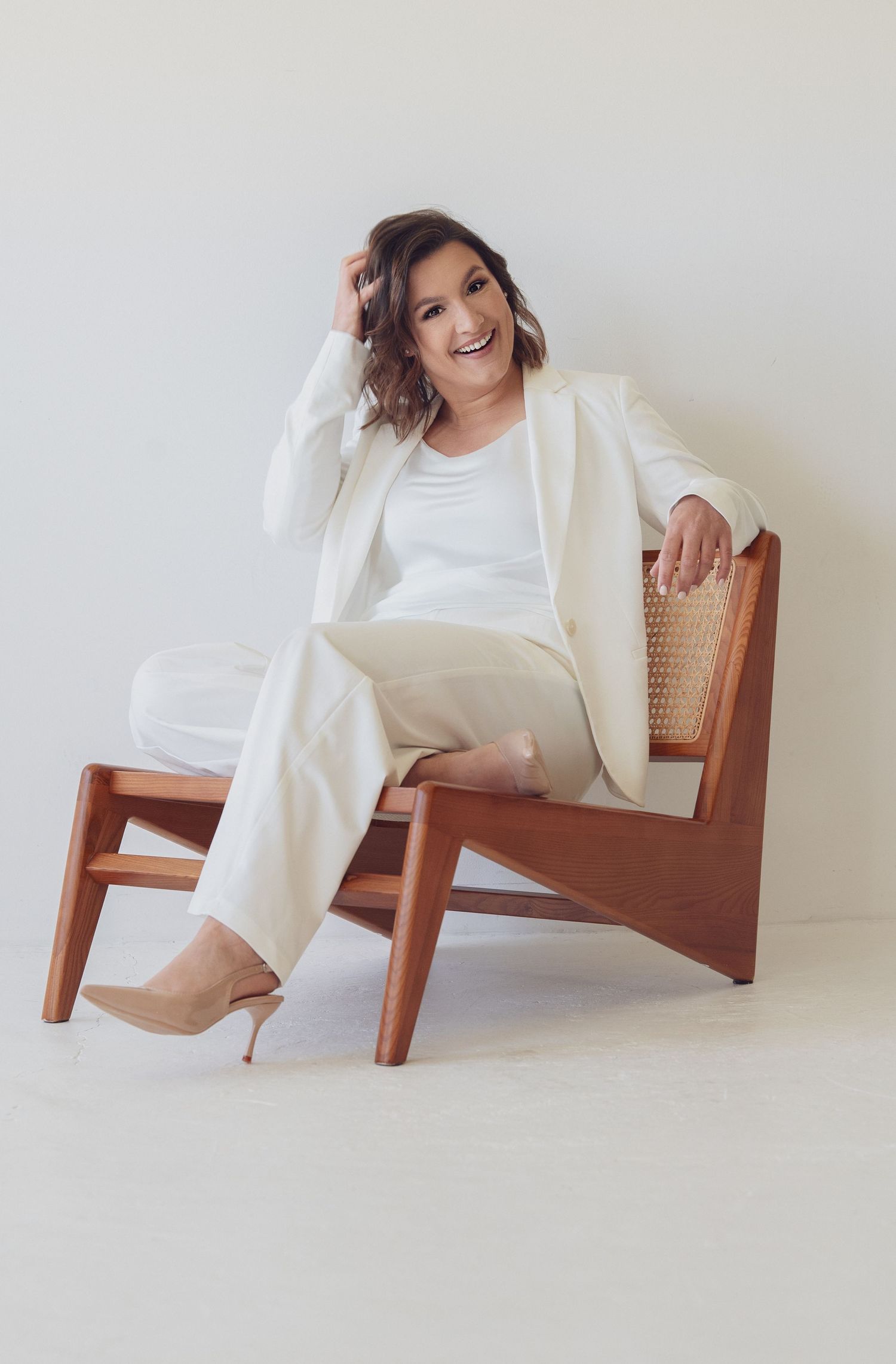 Woman in white blazer and pants sitting relaxed on a wooden rattan chair, smiling confidently in a bright studio.