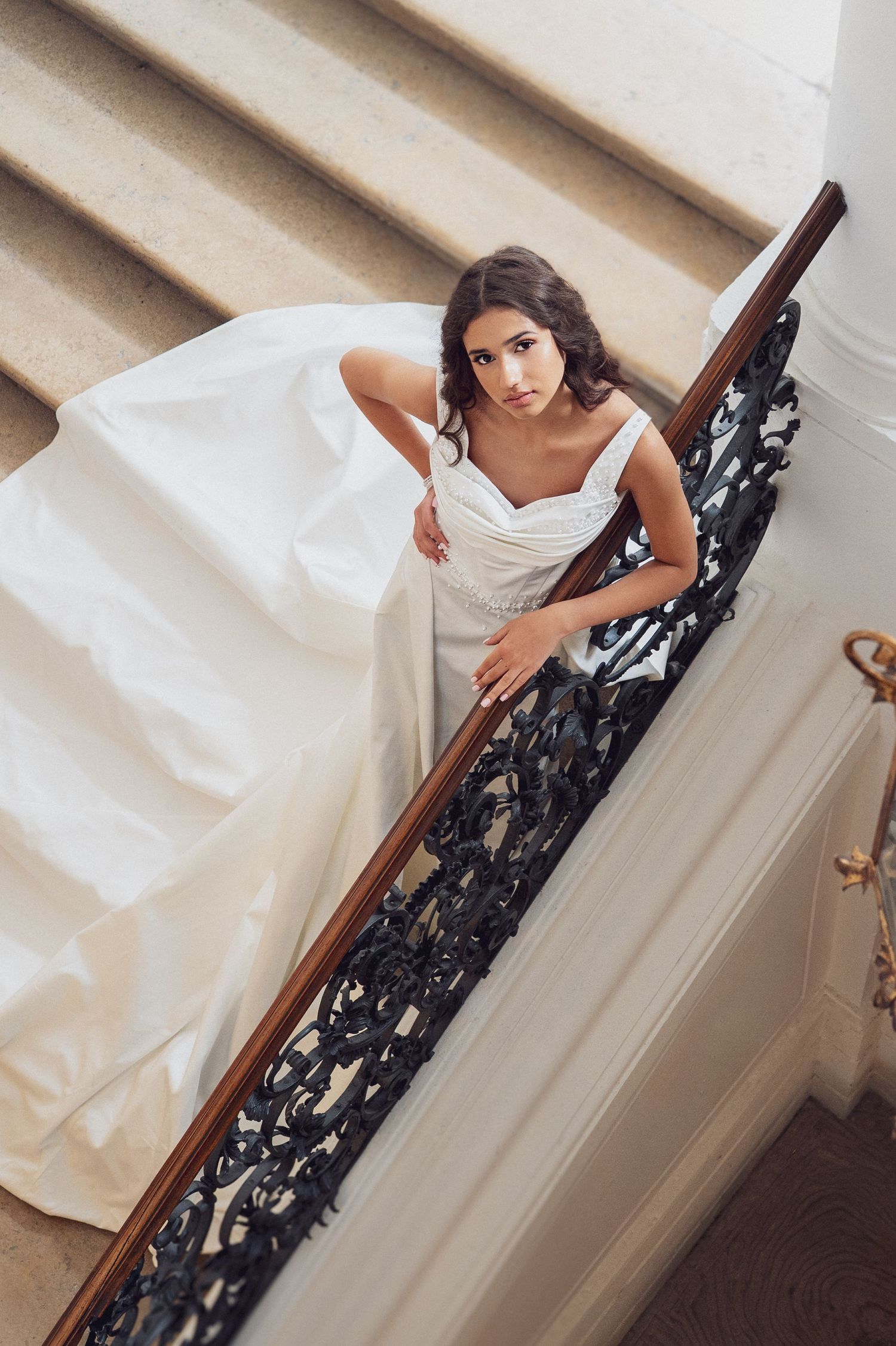 Elegant bride in white ball gown descending ornate staircase with decorative iron railing in grand venue.