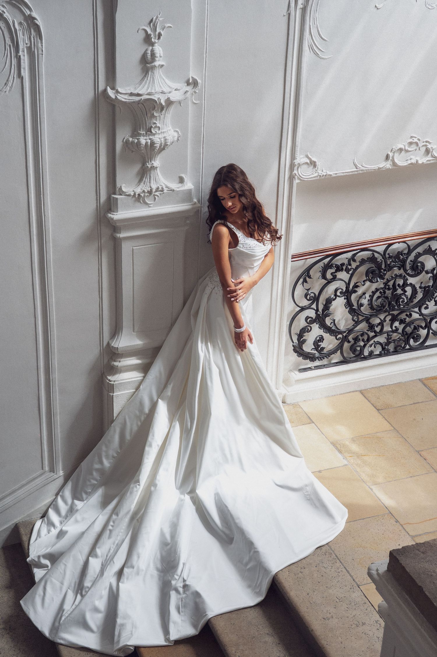 Bride in elegant white satin ball gown with long train poses gracefully on stairs in ornate classical interior.