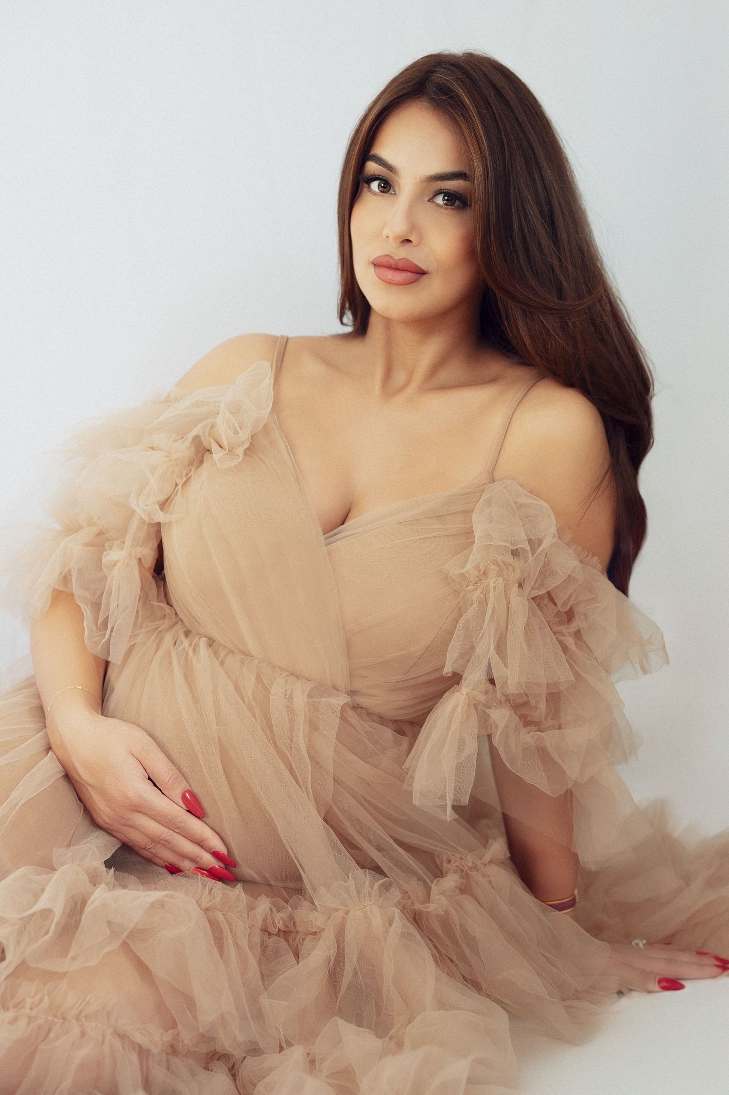 Elegant maternity photoshoot with flowing beige tulle dress against light background.