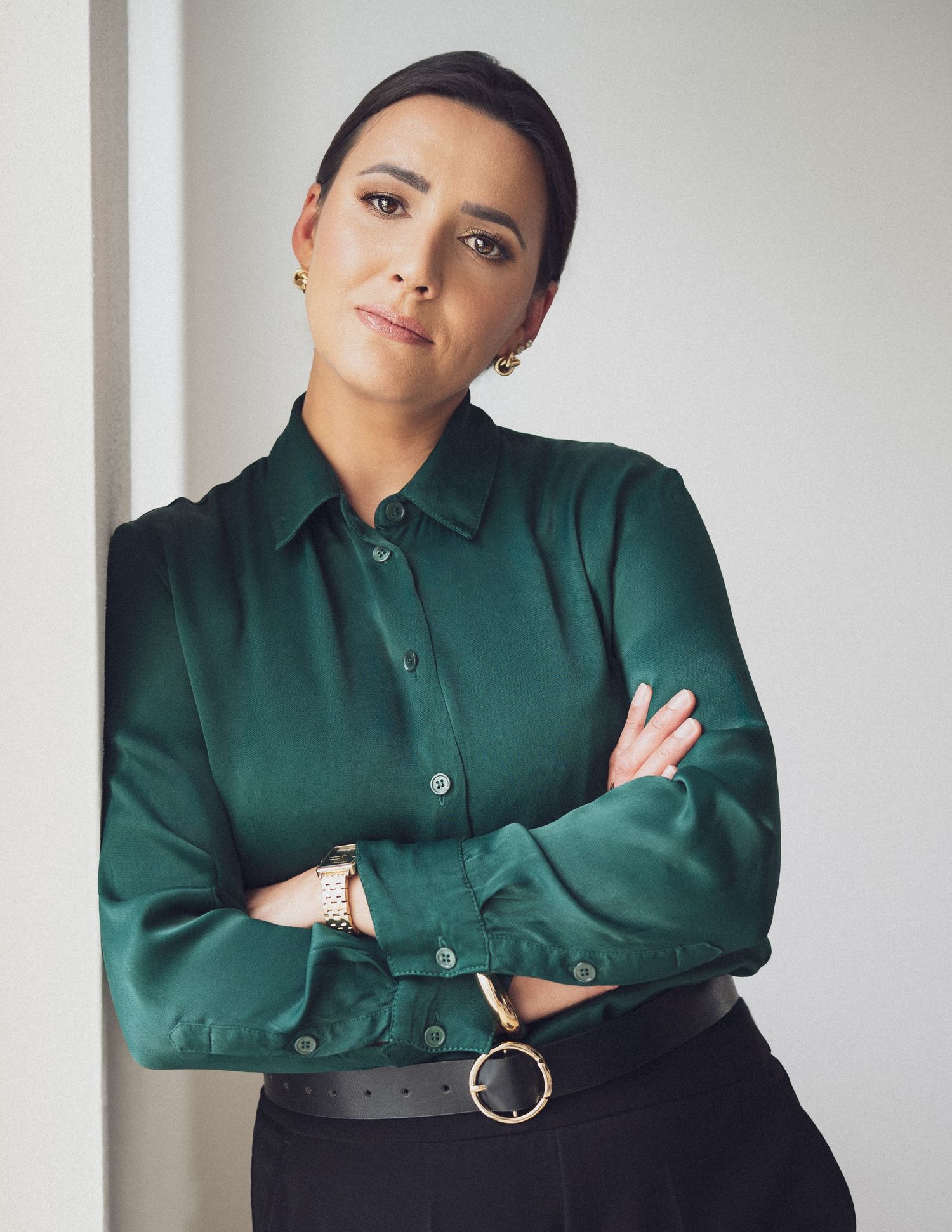 Professional woman in green satin shirt with arms crossed, wearing gold jewelry and belt, posed against white wall.