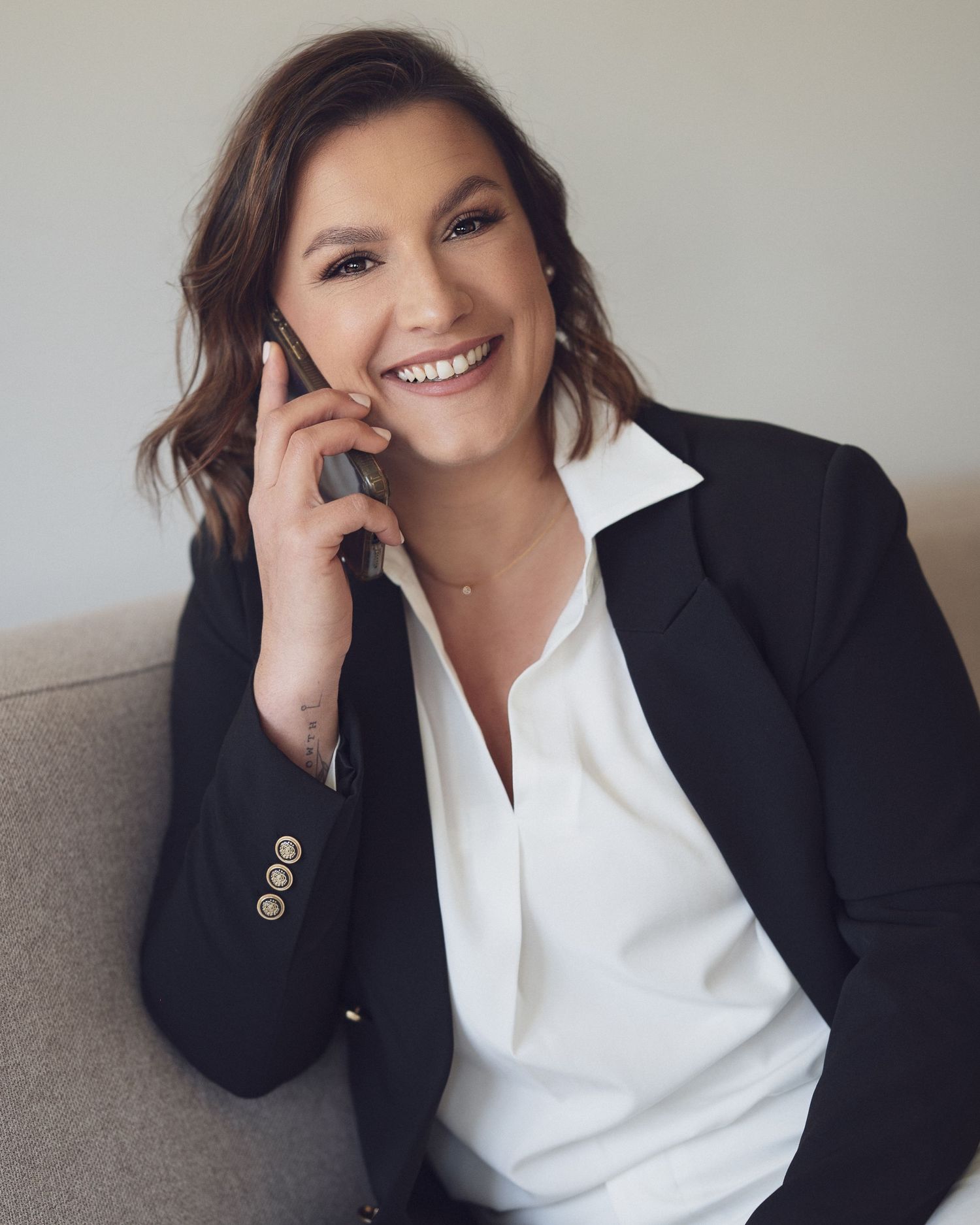 A smiling professional woman in a black blazer and white blouse talks on a phone while seated on a gray sofa.