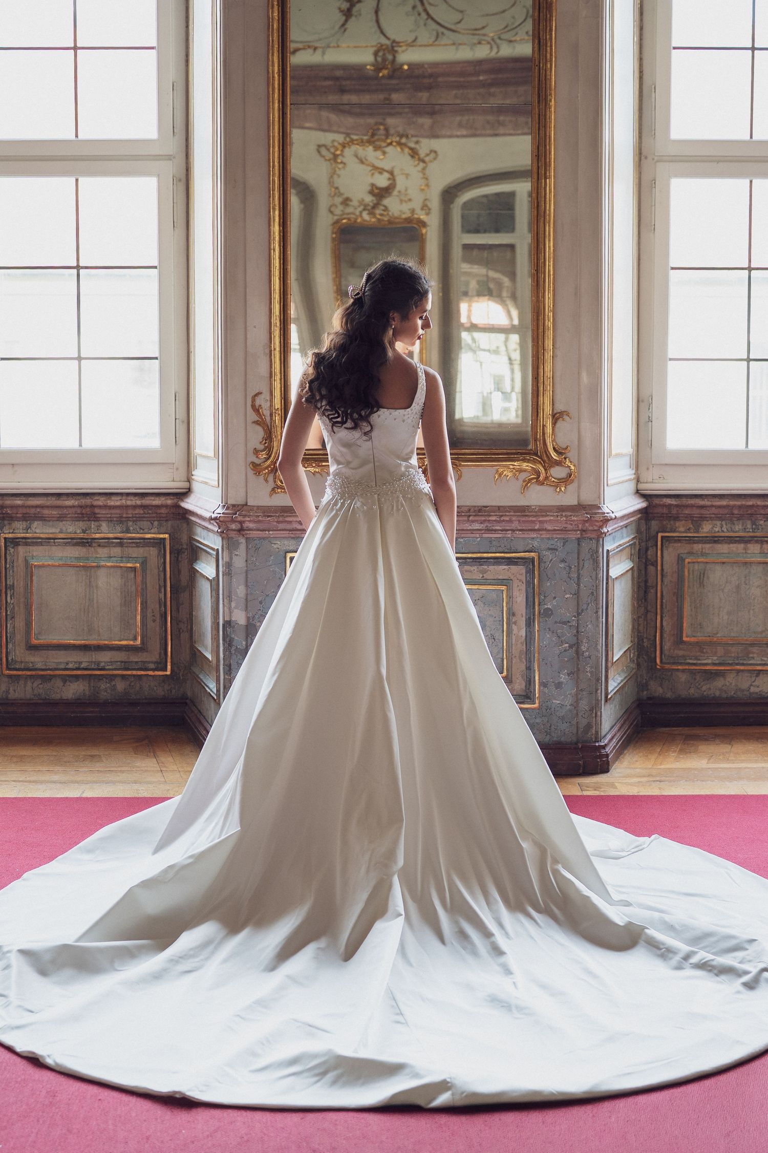 Bride in stunning white satin wedding gown with cathedral train stands before gilded mirror in luxurious ballroom.