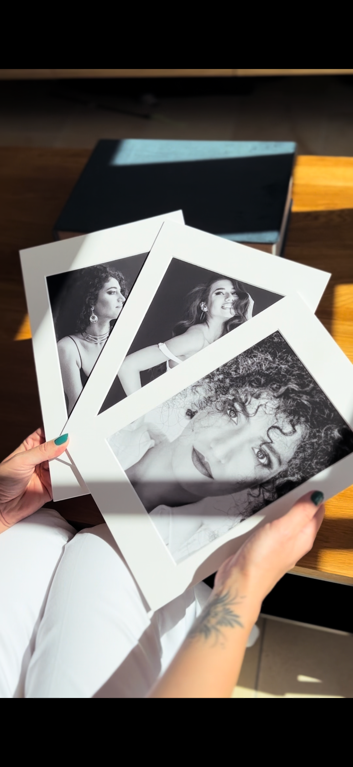 Multiple black and white photographic prints displayed in hands against a dark background with natural lighting.