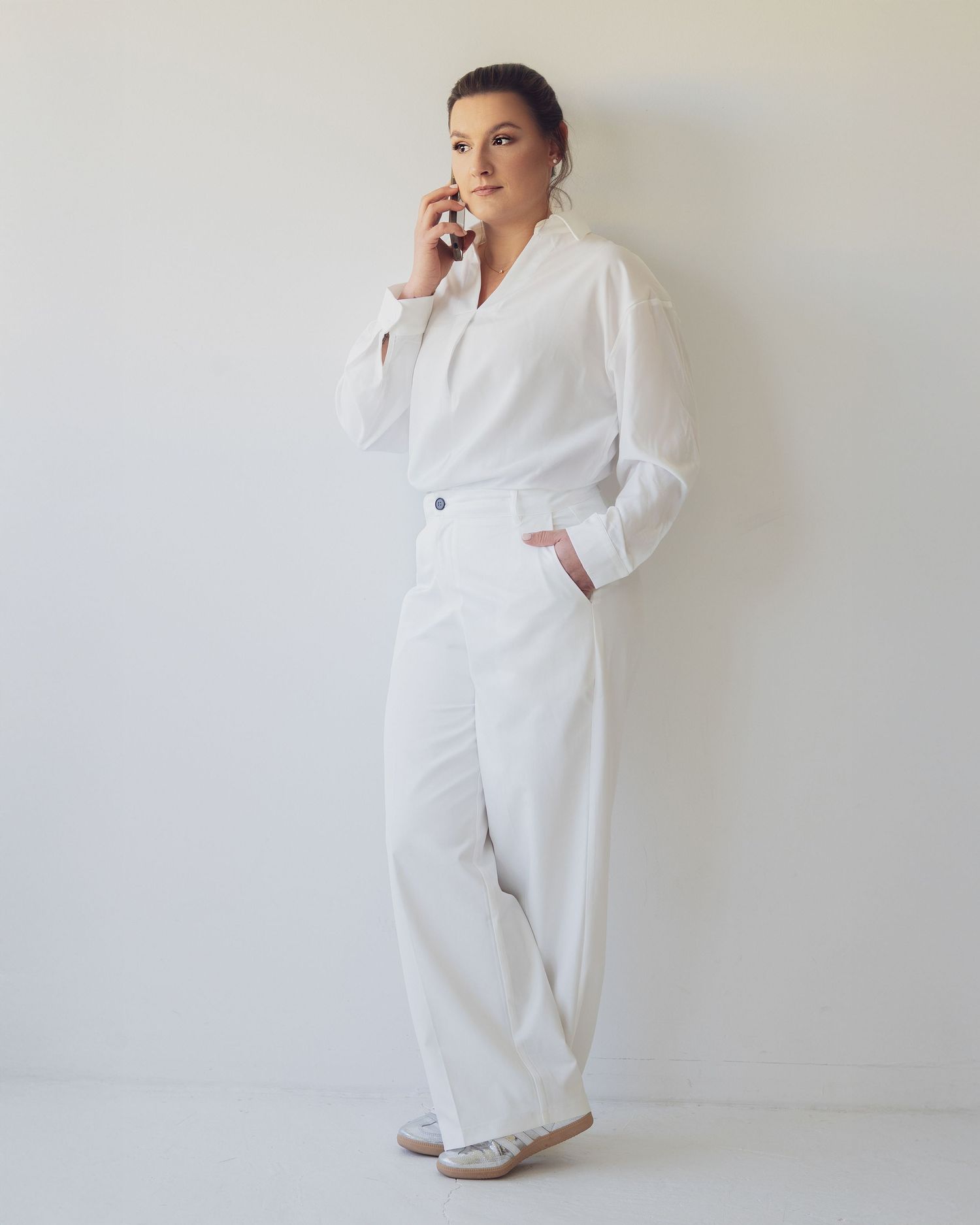 Elegant woman in white outfit talking on phone, standing against light background.
