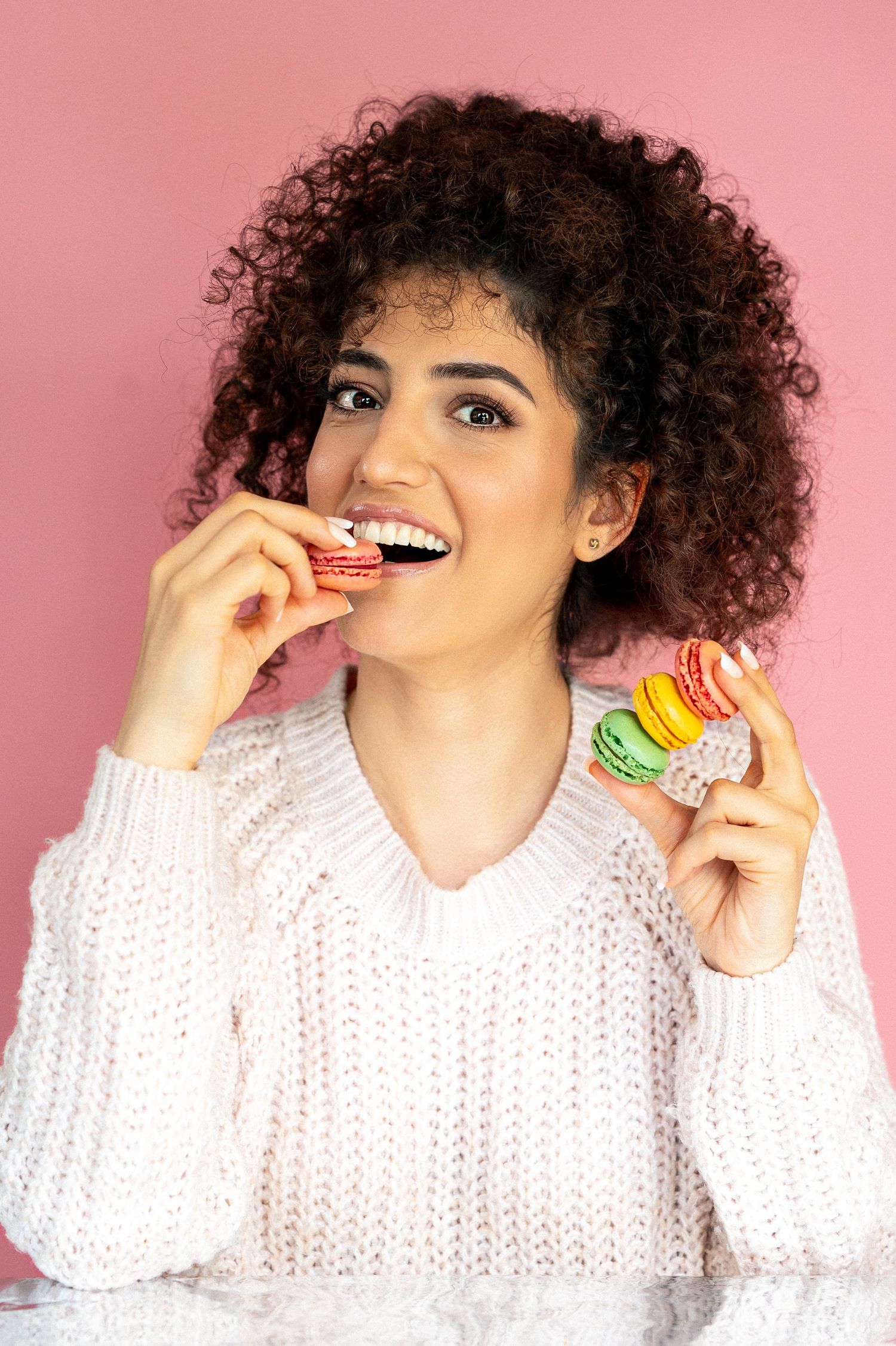 Young woman with curly hair biting into a colorful macaron while holding more macarons, pink background.