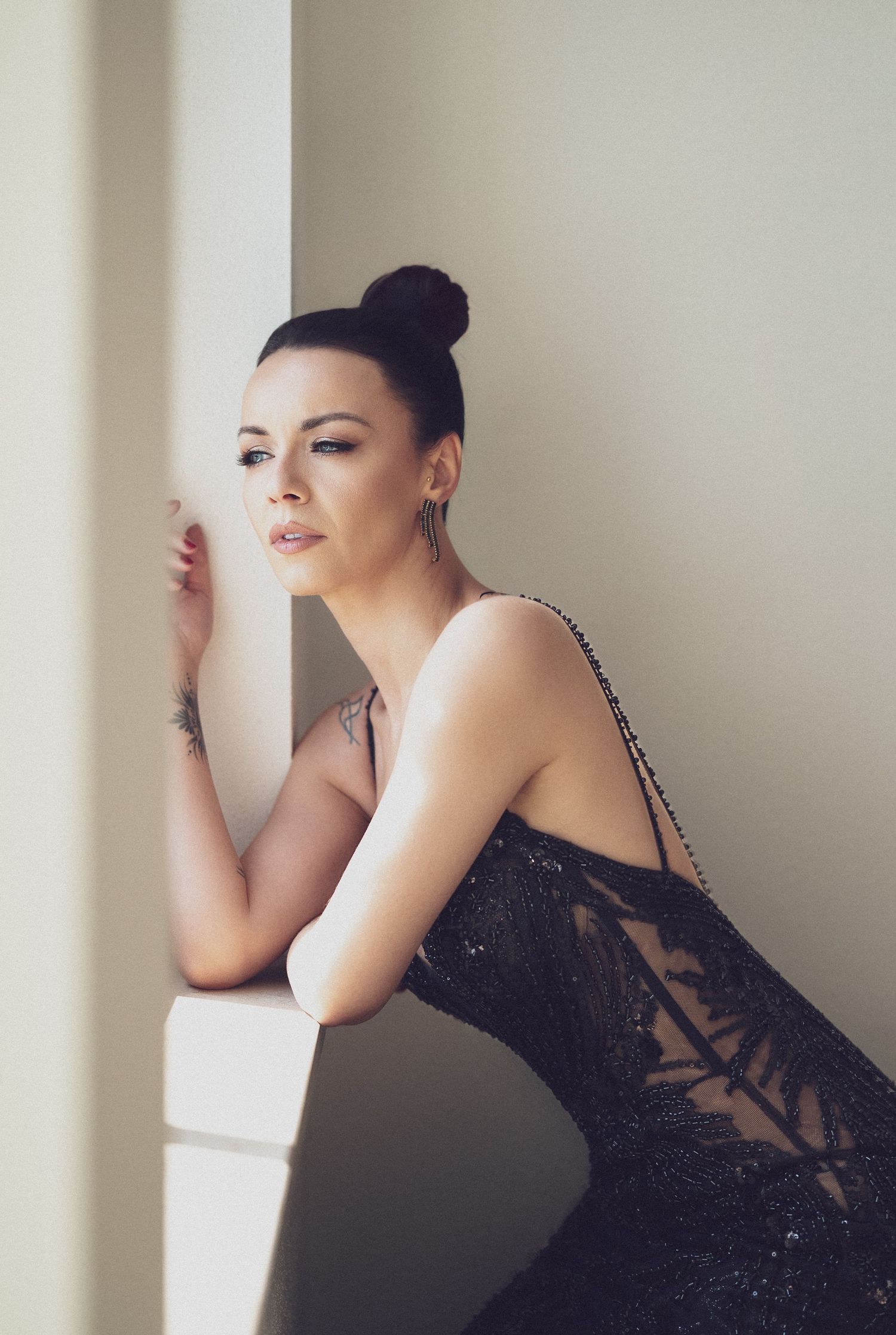 Woman in black lace dress with hair bun leans gracefully against white wall in elegant fashion portrait.