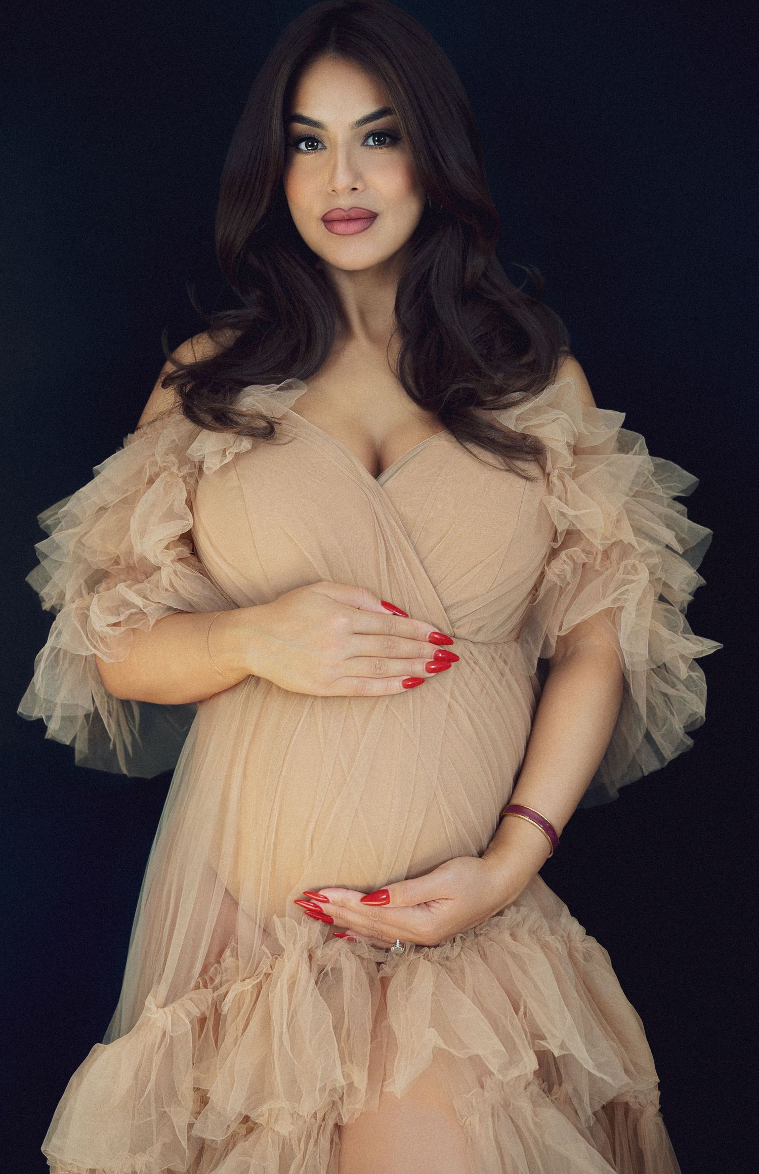 Maternity portrait in peach ruffled dress against dark background with glamorous styling.