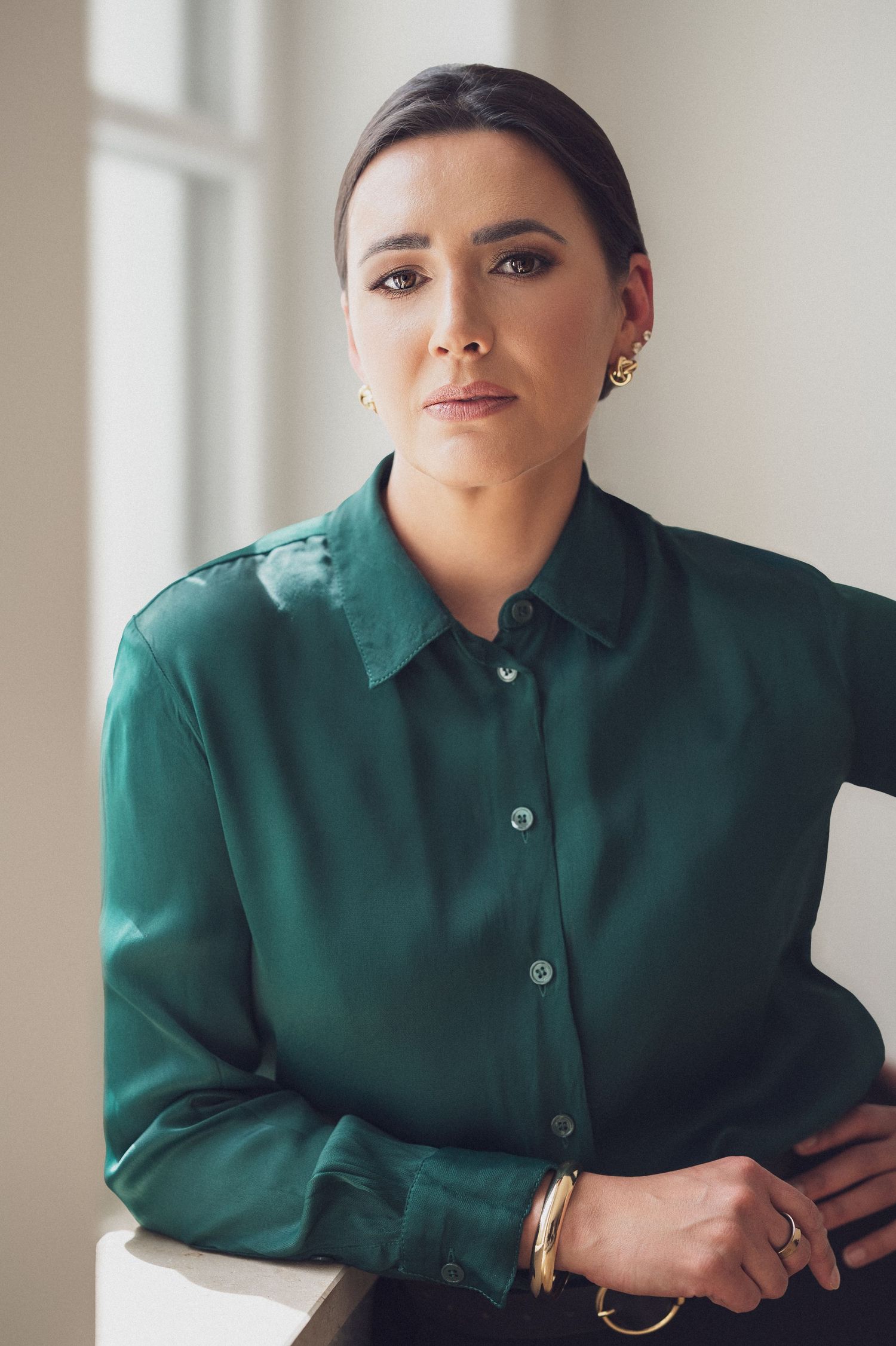 Woman in green silk blouse with gold jewelry poses confidently near a window in a professional portrait.