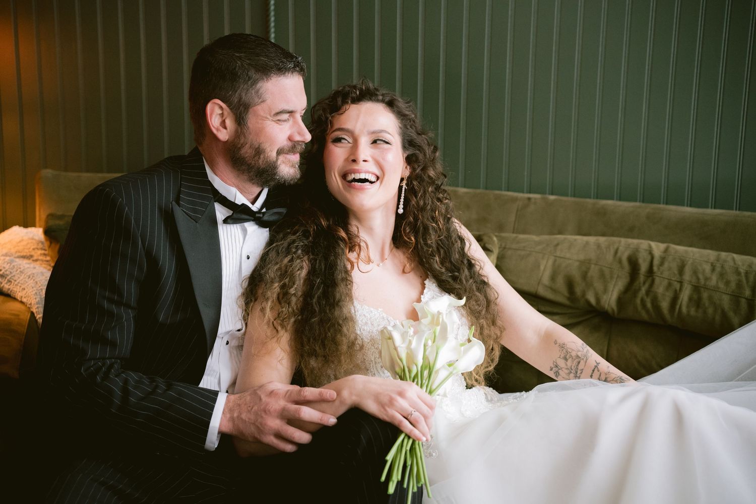 A bride and groom posing on a velvet couch and laughing