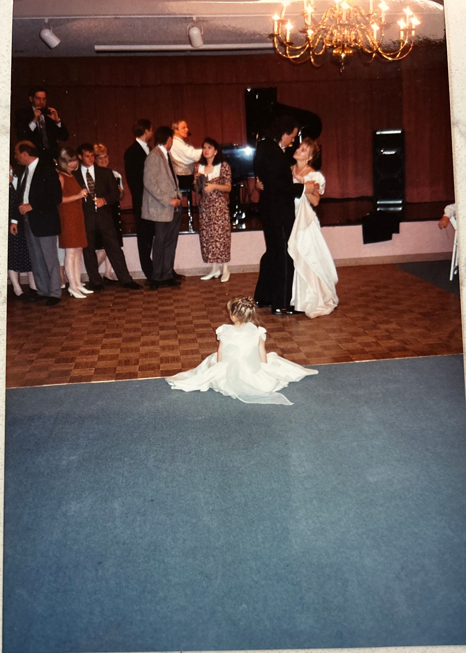 Guests dancing at a formal event in a ballroom with a chandelier and patterned carpet.