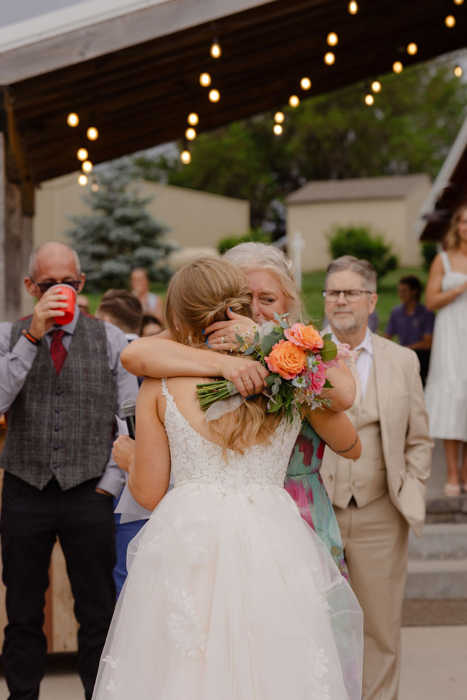 A bride in a white dress hugs someone while holding a bouquet at an outdoor reception.