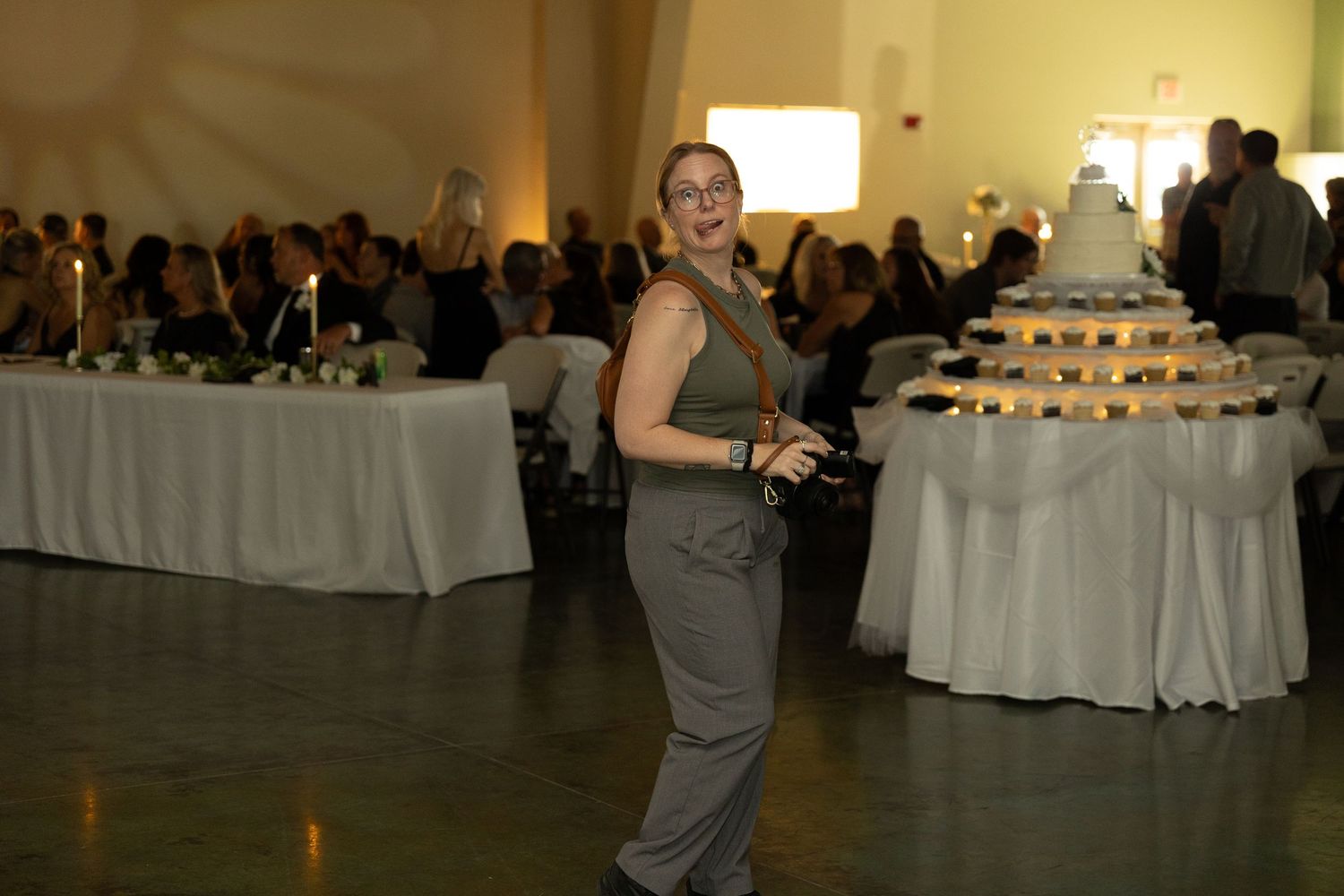 A person in gray clothing stands near a wedding reception dessert table with tiered displays and white tablecloths.