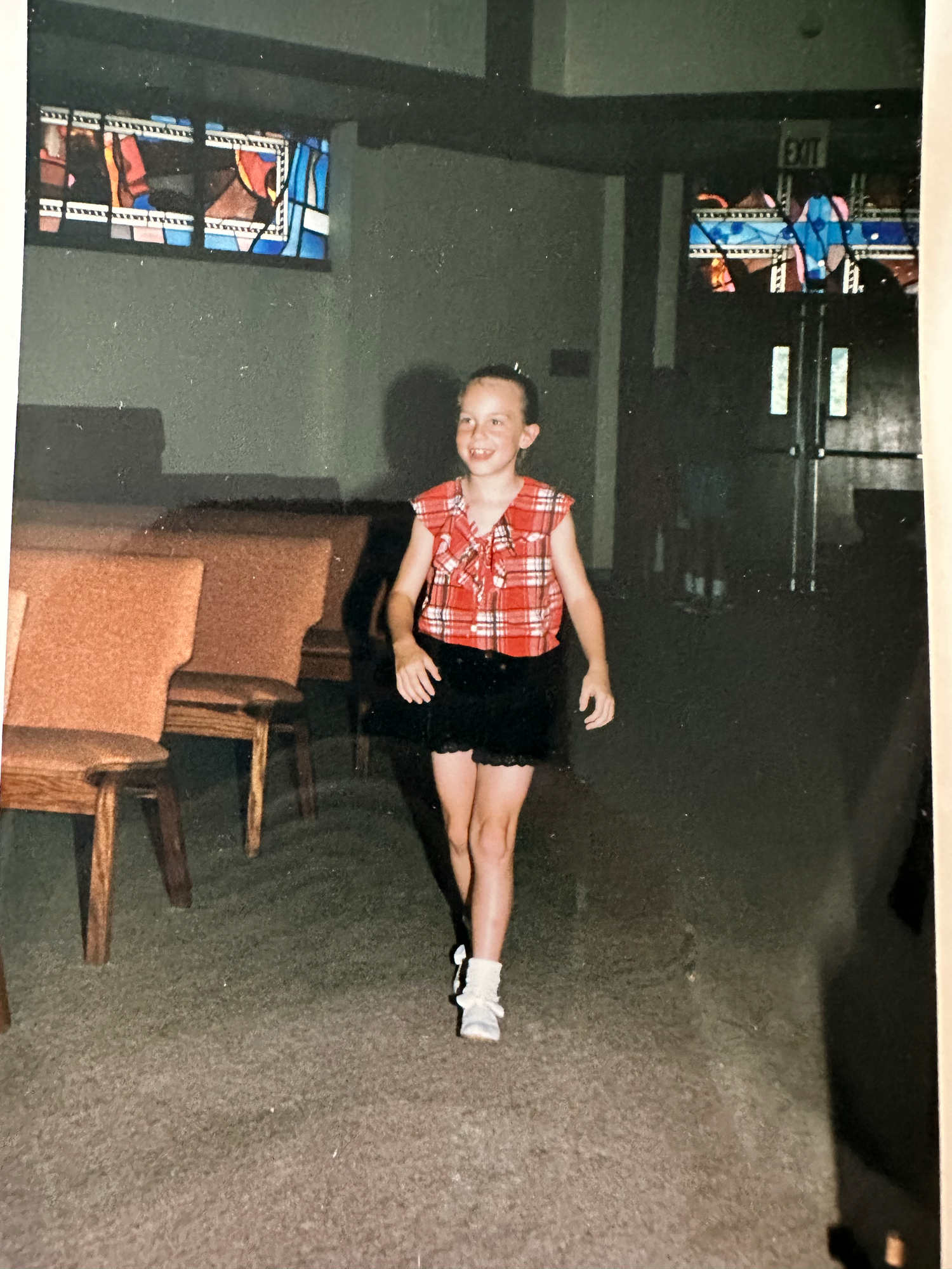 Portrait of someone wearing a red plaid vest and black shorts standing in a dim room with chairs against the wall.