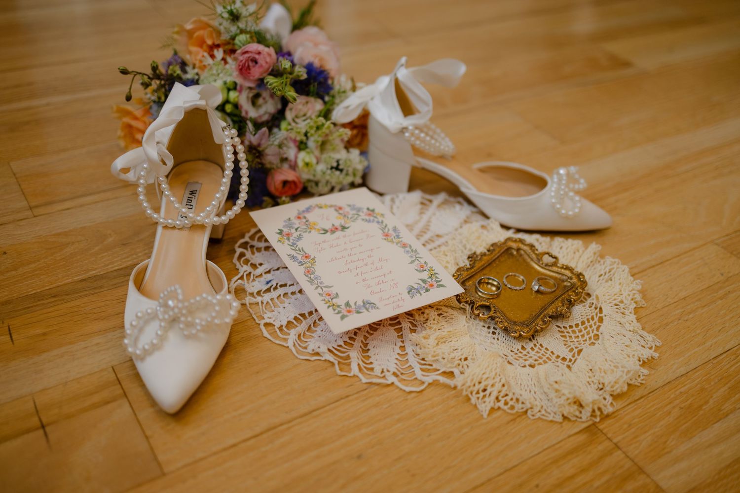 Wedding bouquet with pink and purple flowers wrapped in white lace and ribbon rests on wooden floor. Arbor Hall, Omaha Nebraska