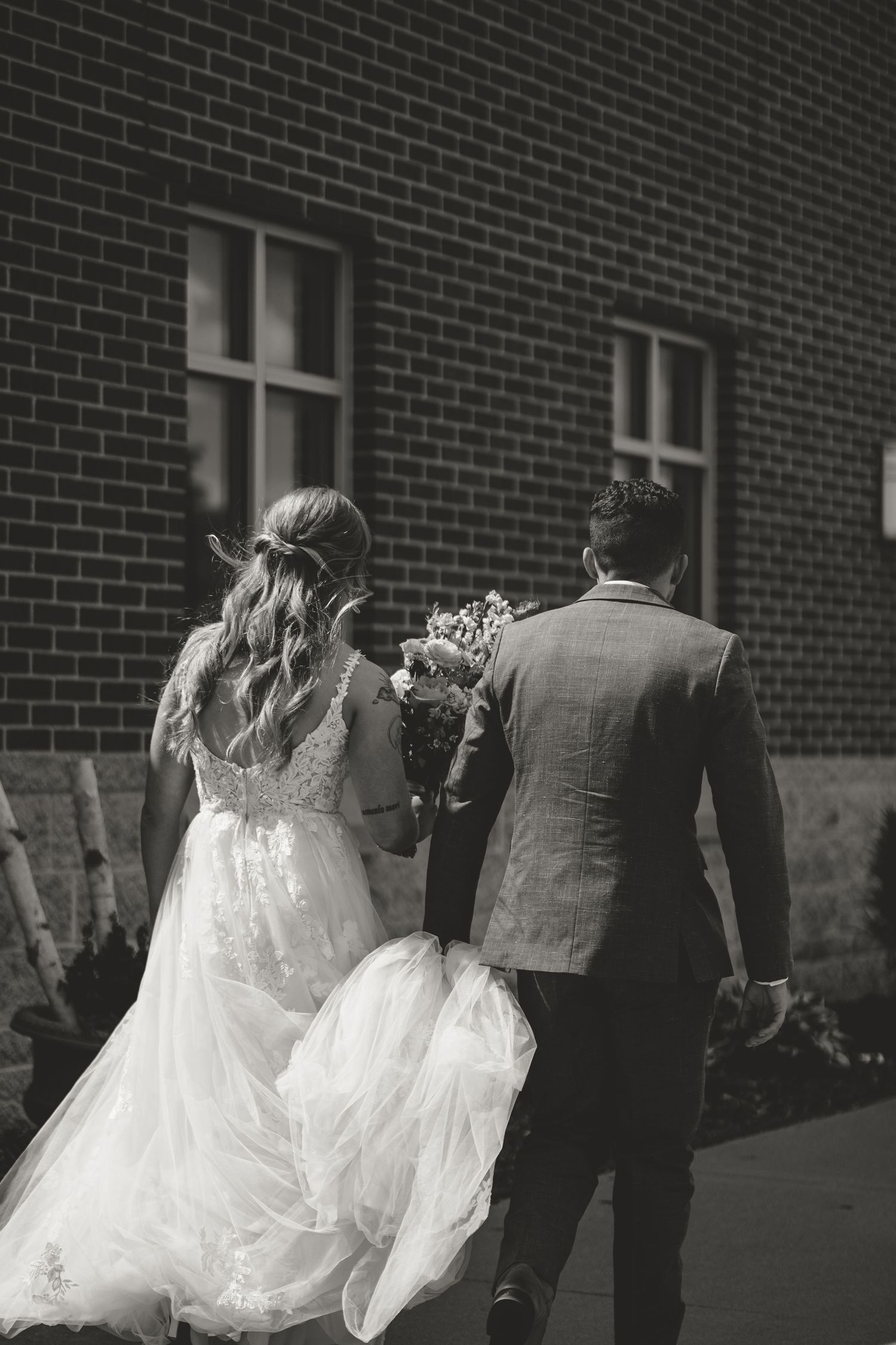 Bride and groom walk together along a brick building in black and white, holding hands with flowing wedding dress trailing behind.