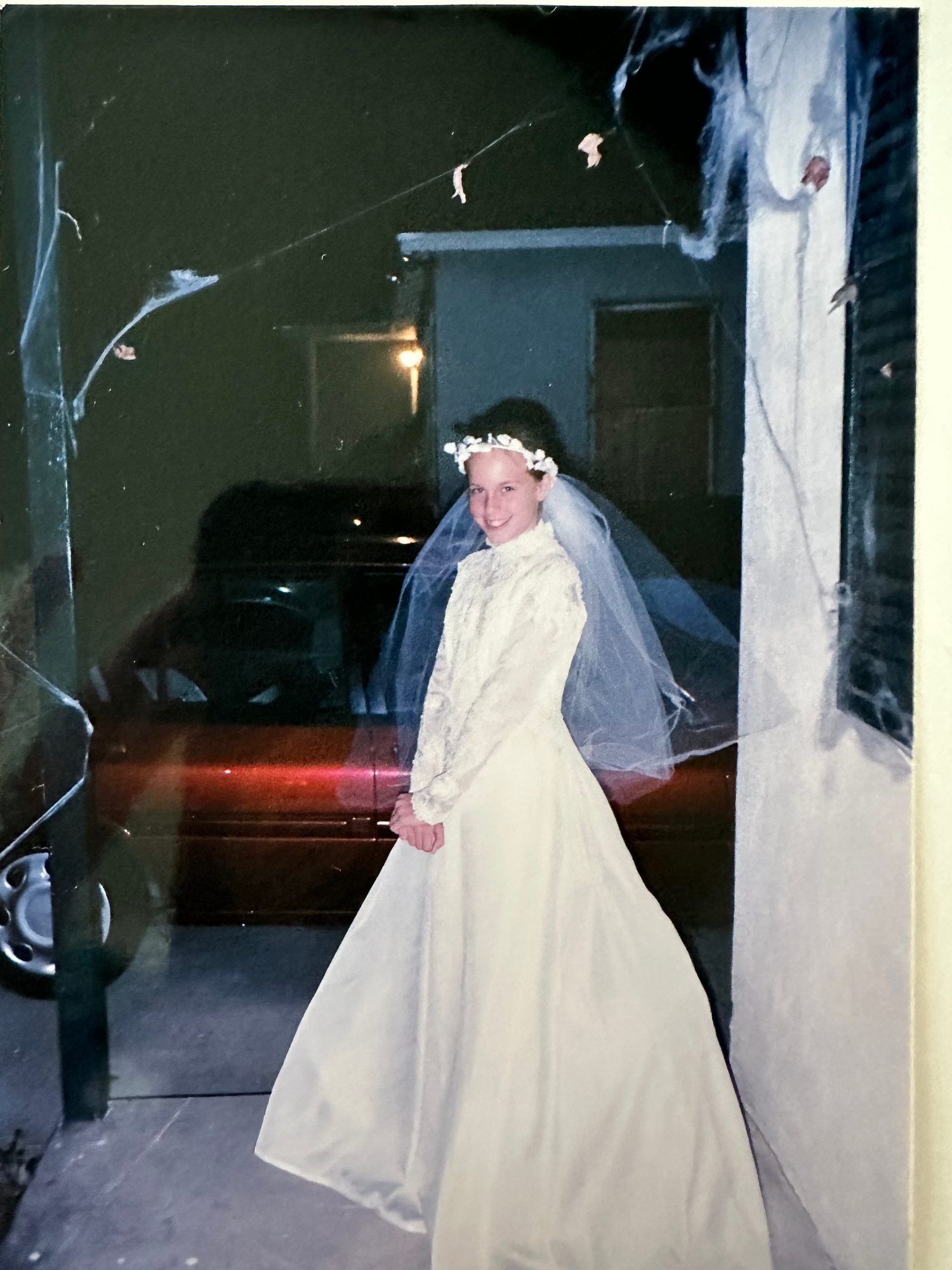 A bride in a classic white wedding gown and veil stands in front of a car at night with outdoor lighting visible.