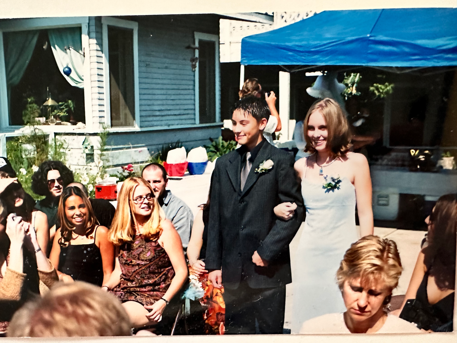 A group stands together at an outdoor wedding ceremony under a blue canopy in warm sunlight.