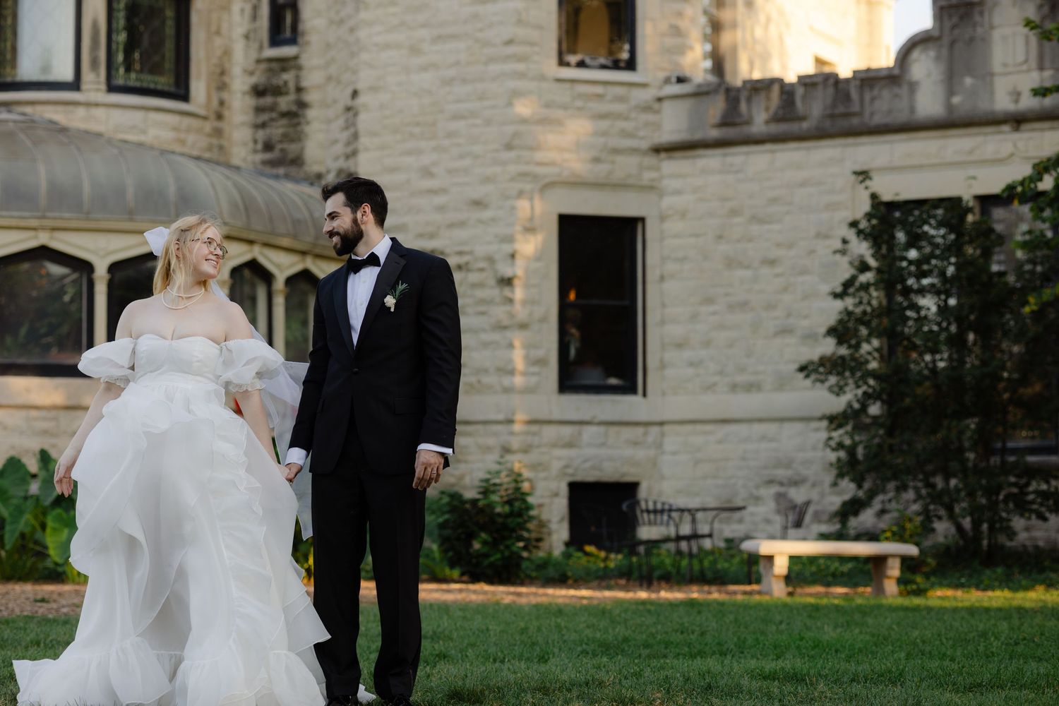 A bride and groom share a romantic moment together on the grounds of an elegant stone estate at sunset.
