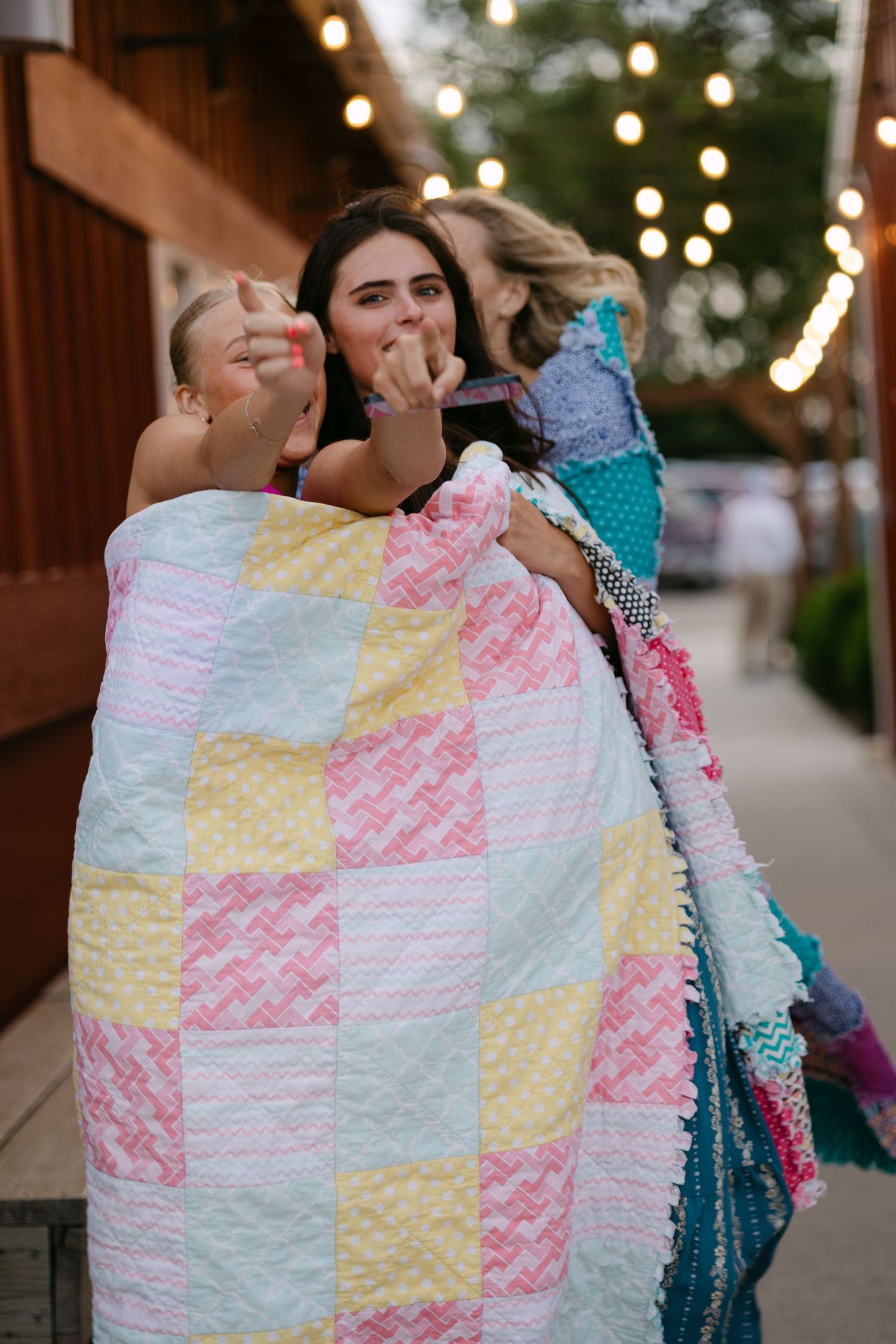 Two cozy friends sitting together wrapped in colorful patchwork quilts with string lights glowing in the evening background.