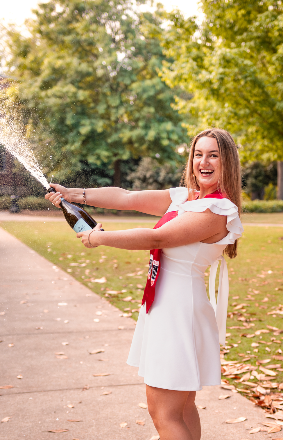 Young woman in white dress joyfully popping champagne bottle outdoors on a tree-lined path.