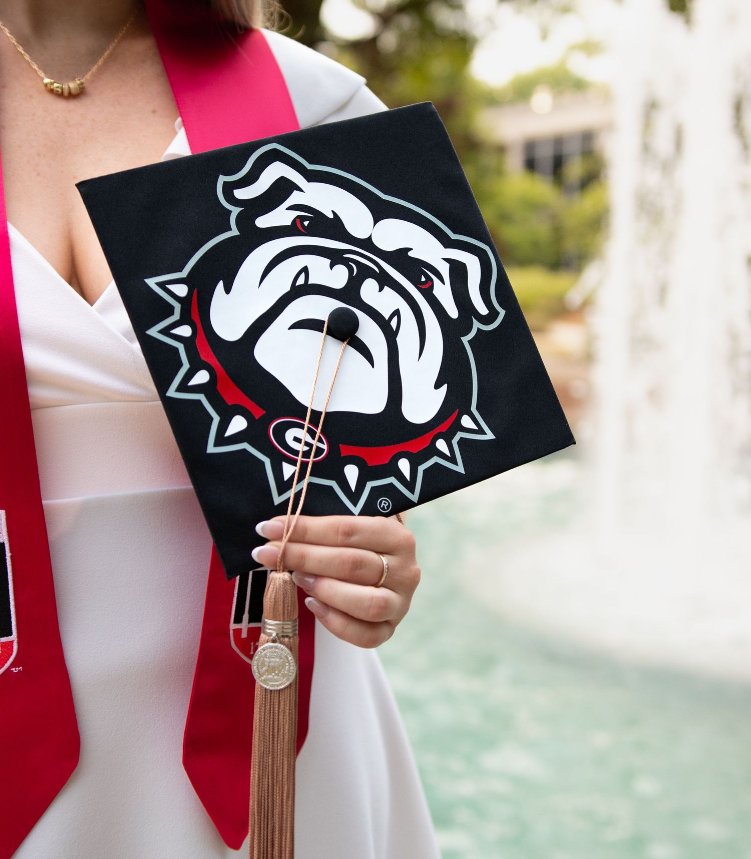Graduate holds a black UGA Bulldogs graduation cap with mascot logo, wearing white gown and red stole.