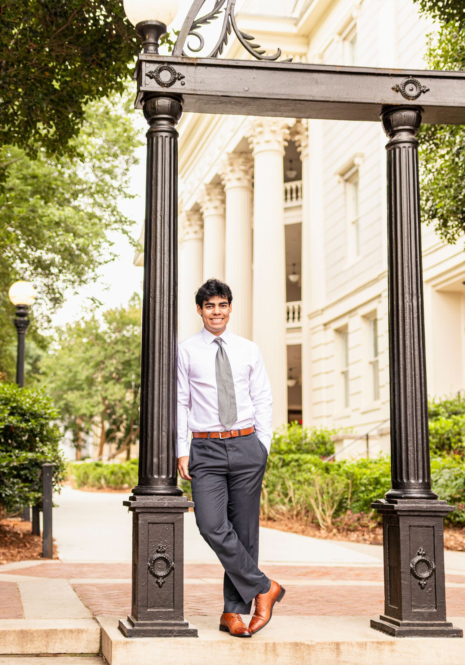 Young man in dress shirt and tie posing between ornate iron columns outside a classical building.