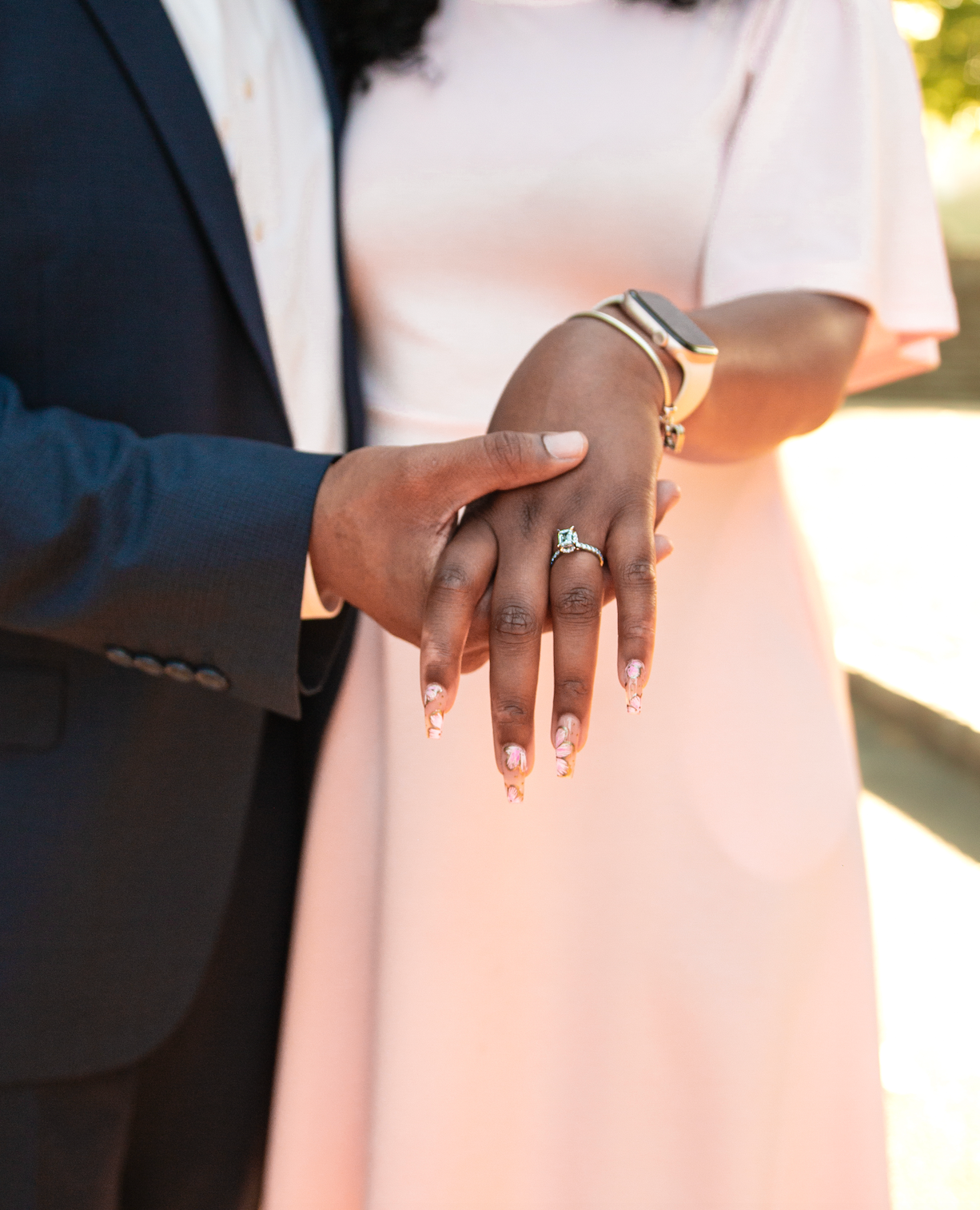 Close-up of a couple holding hands displaying an engagement ring, with the woman wearing a blush pink dress and decorated nails.