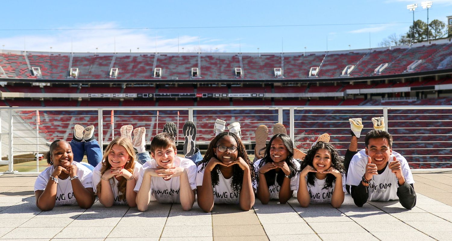 A group of cheerleaders pose lying on their stomachs in a stadium setting during a sunny day.