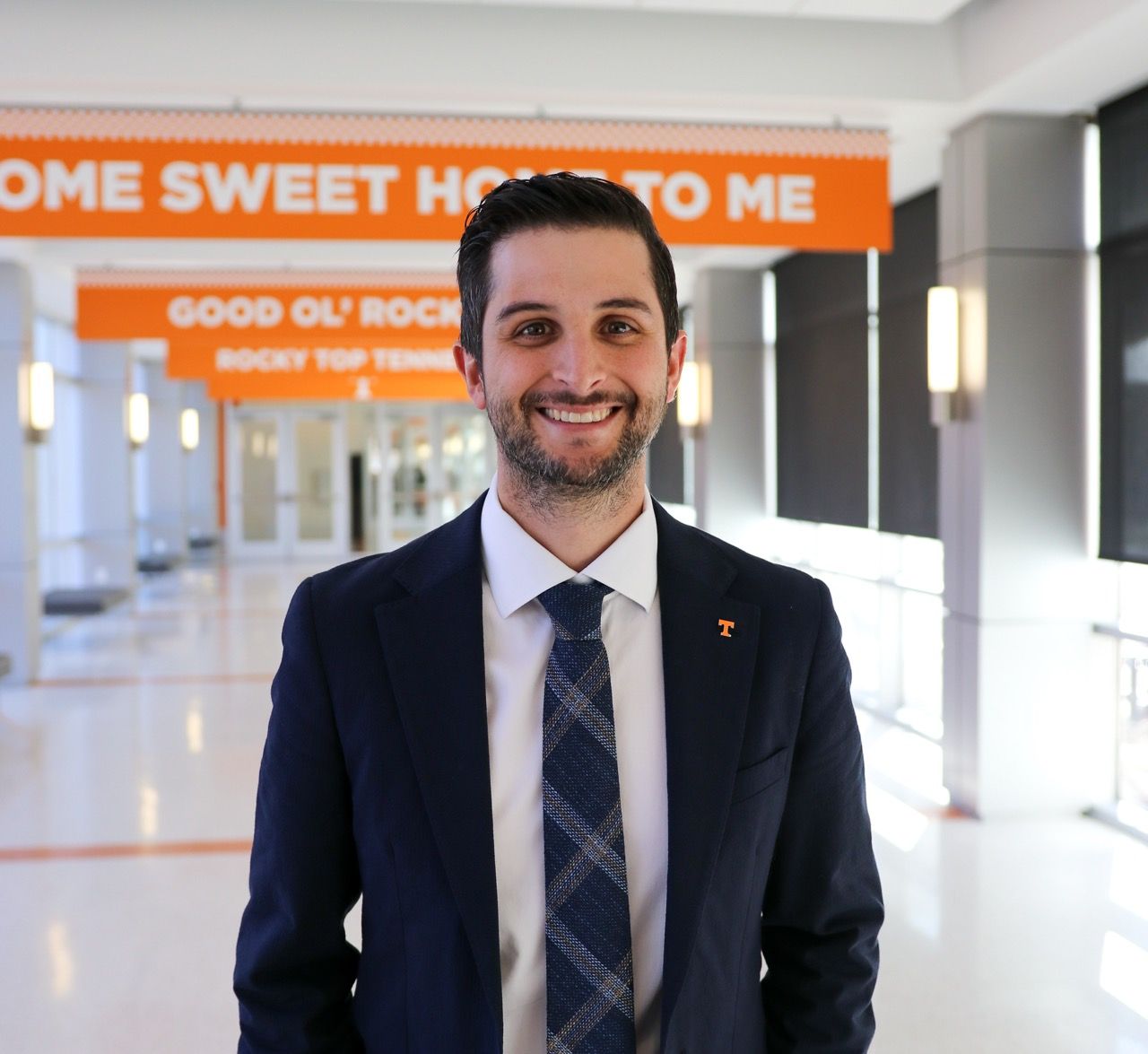 Professional wearing a navy suit and tie stands in a modern office hallway with orange signage visible in background.