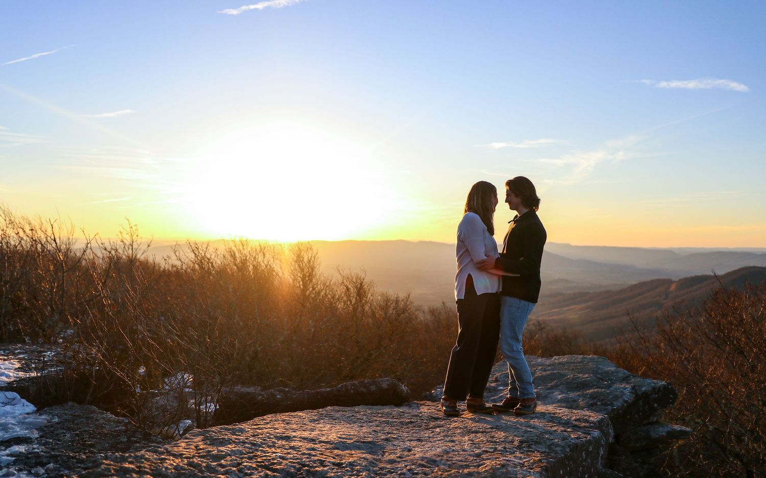 A silhouetted couple embraces on a hilltop against a golden sunset sky with rolling mountains in the distance.