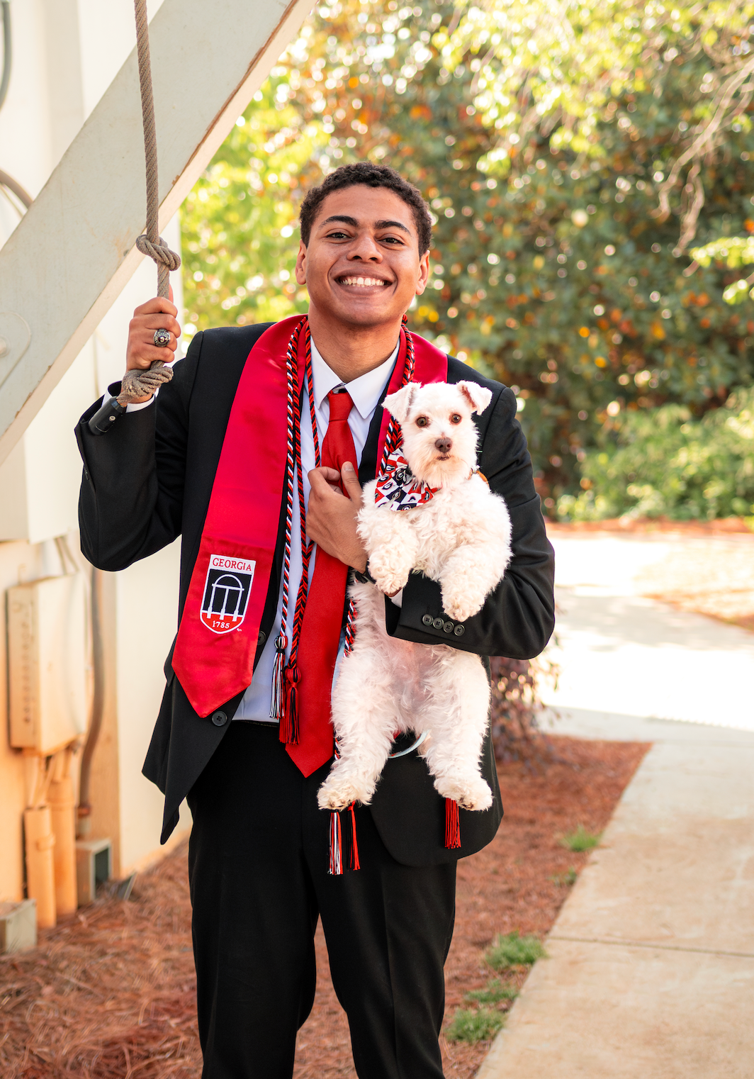 Smiling graduate in black suit with red stole holds white fluffy dog outdoors on graduation day.