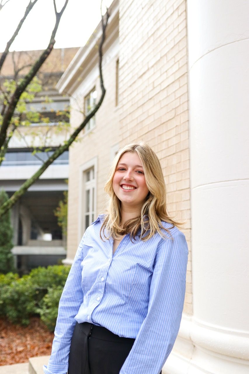 Professional business portrait in light blue striped button-down shirt against brick wall outdoors.