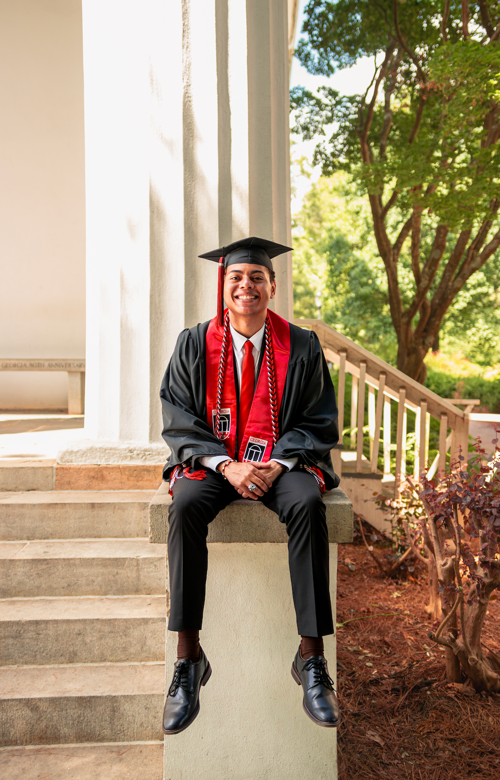 Smiling graduate in black cap and gown with red stole sits on steps by white columns outdoors.