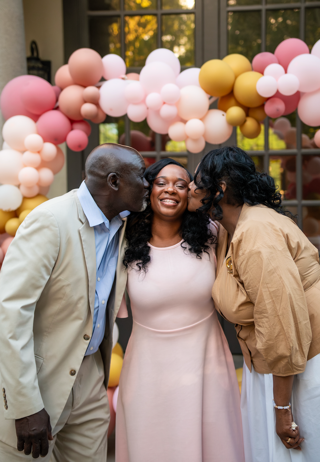 A joyful woman in pink dress receives kisses on cheeks from two loved ones in front of colorful balloon arch.