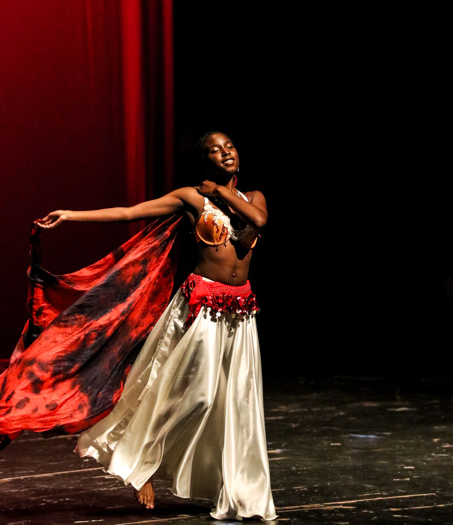 A dancer in a flowing white dress with red fabric performs an expressive movement sequence on a dark stage.