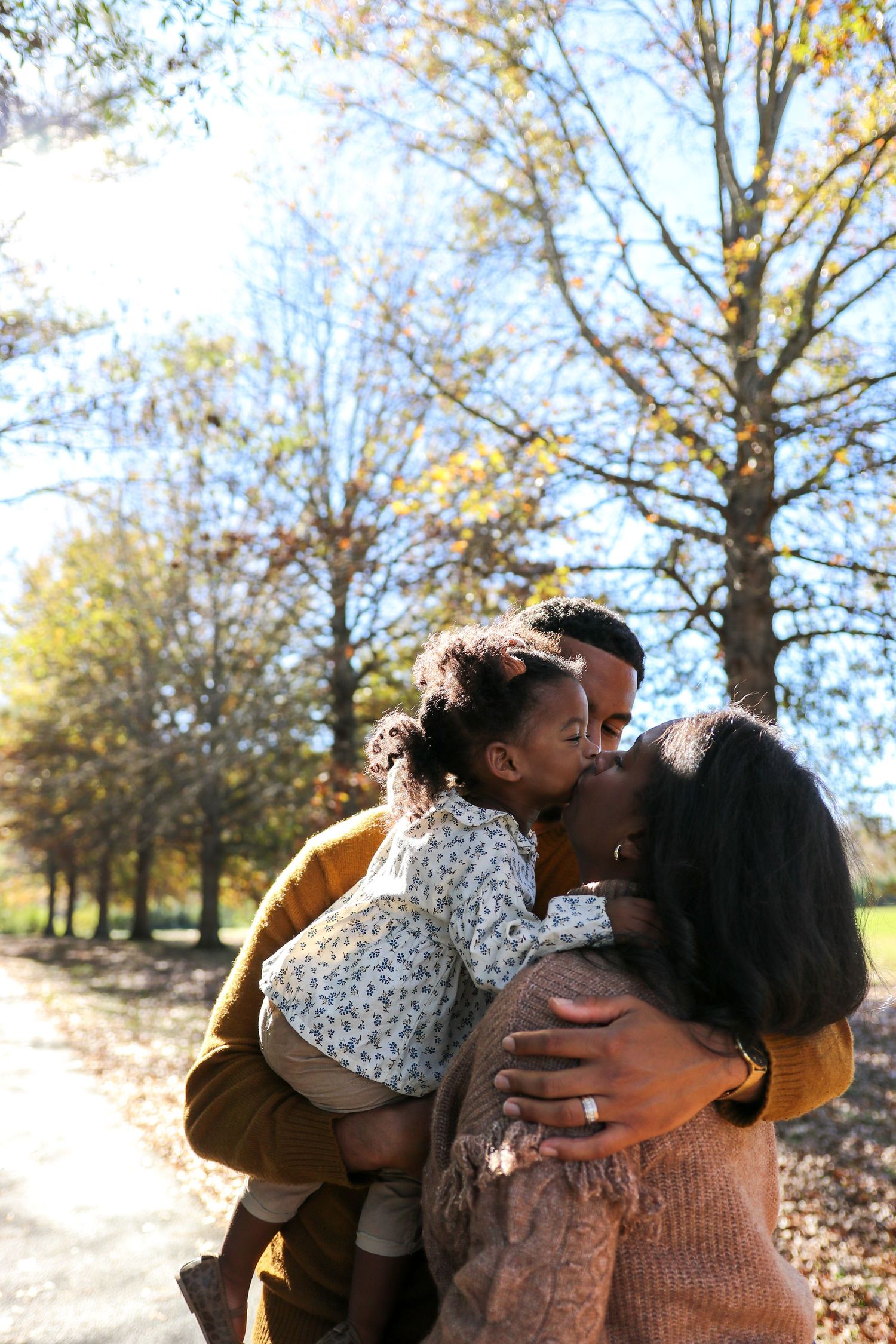 A couple embraces and kisses while surrounded by autumn trees and fallen leaves in a park setting.