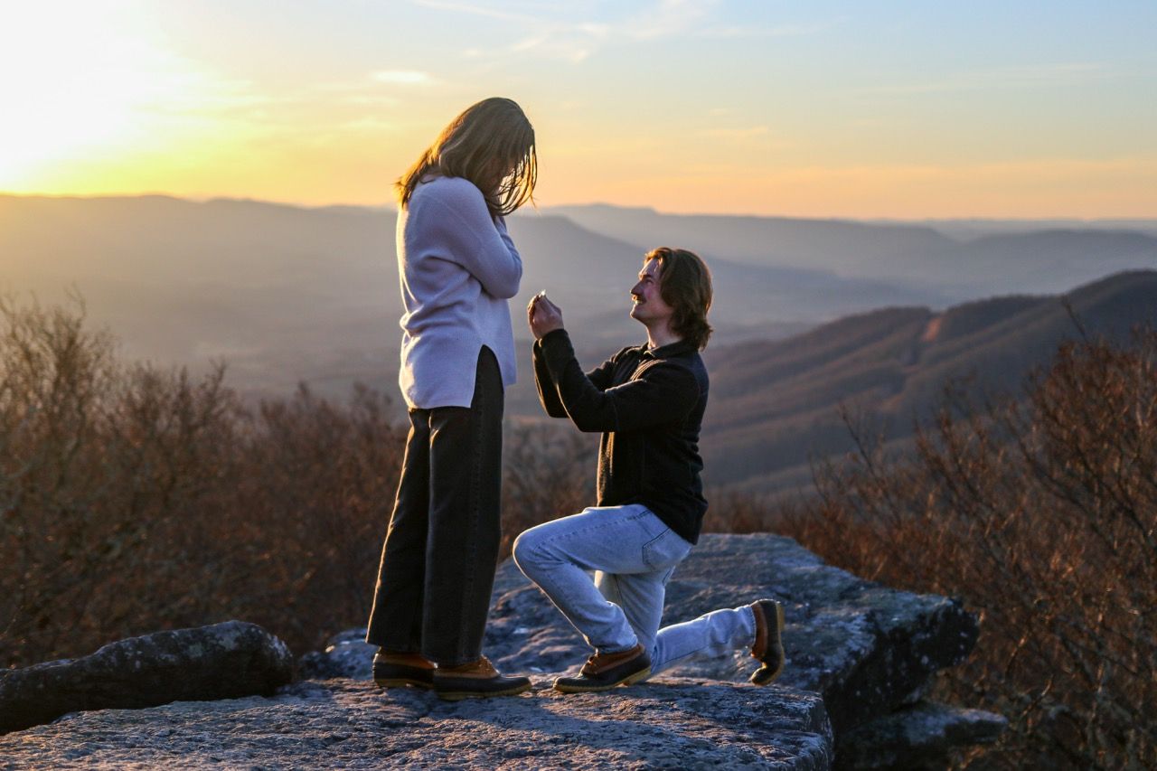 A couple enjoys a scenic mountain overlook at sunset while having a picnic with wine and snacks.