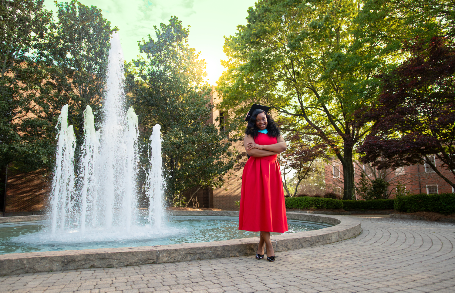Woman in red dress poses confidently by a fountain on a sunny campus with lush green trees.